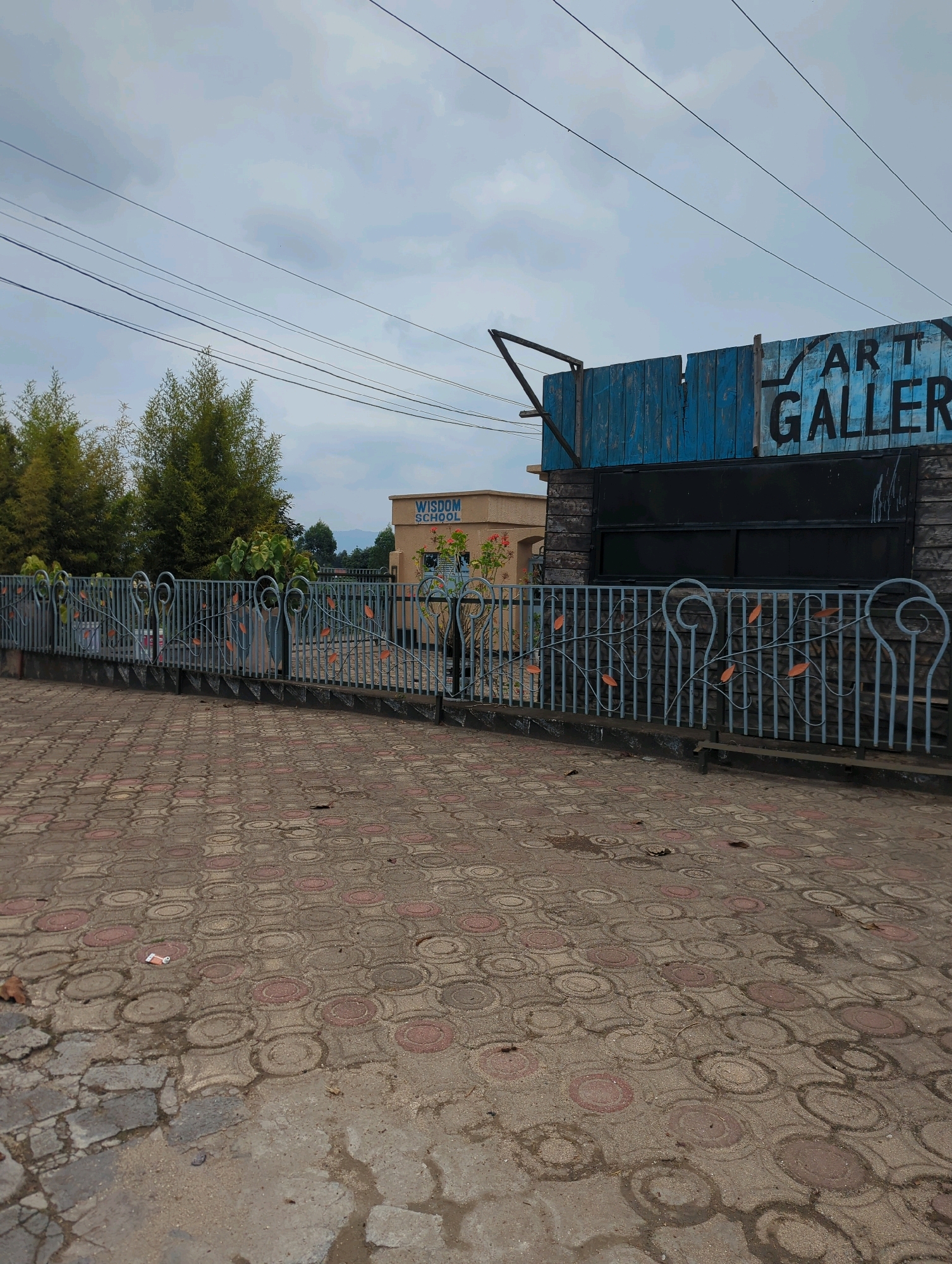In Musanze District, Rwanda, a street scene unfolds under a cloudy sky. A paved walkway, made of circular patterned interlock bricks, dominates the foreground. A grey metal fence with decorative swirls and leaf motifs separates the walkway from a property. Beyond the fence, a building with a light-colored facade is visible, displaying the sign "WISDOM SCHOOL" in black lettering. Further to the right, a blue wooden structure with peeling paint bears the black lettering "ART GALLERY" on its upper section. A large, dark, rectangular window or entrance is set into the lower part of this structure. Several power lines stretch across the sky above. Trees with dense green foliage are situated behind the fence, to the left of the school building.
