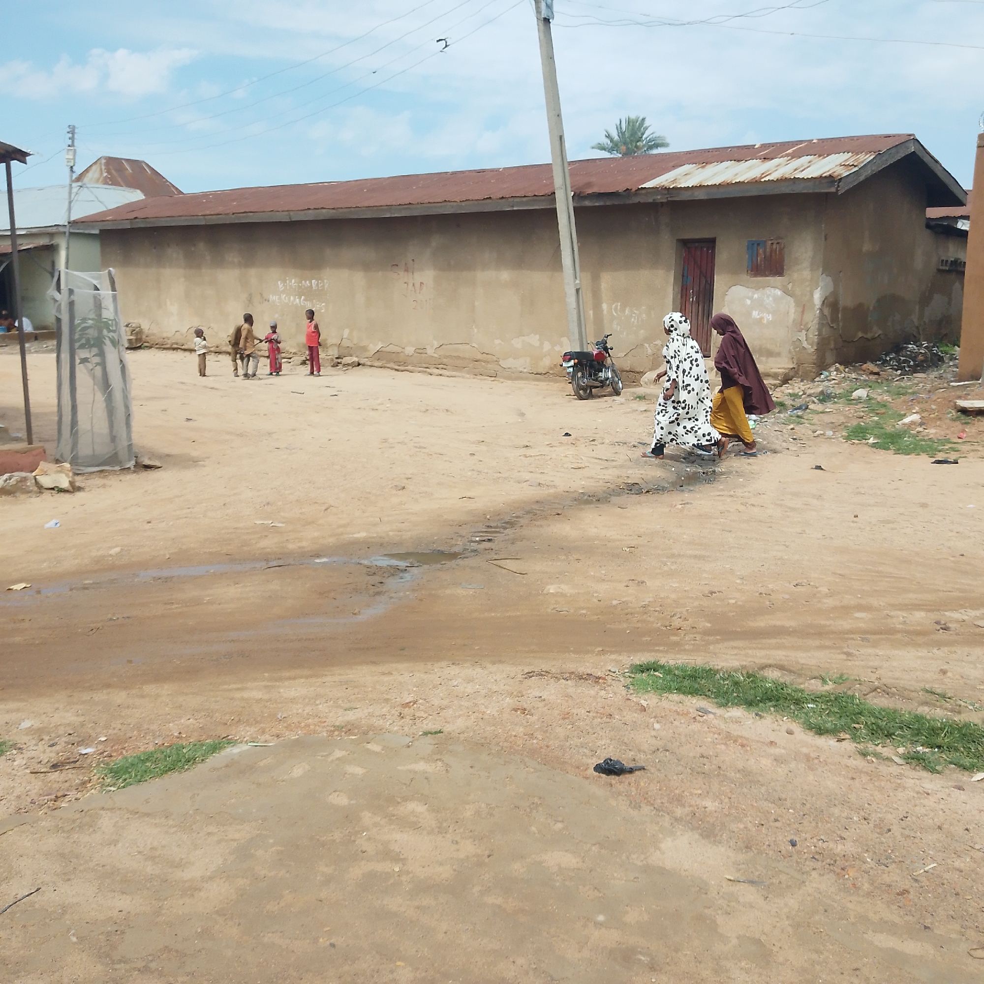 The image depicts an outdoor scene in a dusty, possibly rural or undeveloped urban area. A long, single-story building with a corrugated metal roof and plastered walls dominates the background. The building shows signs of wear and tear, with some discoloration and cracking on the walls. There are two doors visible, one red and one with a blue frame, and a small, barred window.
In the middle ground, a group of four children, two boys and two girls, are gathered. They are dressed in simple clothing. One boy in a brown shirt and pants stands behind a younger child in a white top and red shorts. A girl in a red dress stands to the right of the boys, and another child in a red outfit stands further right.
To the right of the children, two women are walking away from the viewer. The woman in front is wearing a white garment with black polka dots, and the woman behind her is wearing a dark brown head covering and a yellow skirt. They are walking on a dirt path.
A red motorcycle is parked near the building to the right of the children. The ground is predominantly dirt and sand, with some puddles of water and scattered debris. Some sparse green grass is visible along the edge of the dirt path in the lower right of the image. A tall, concrete utility pole with wires stretches across the upper portion of the scene. In the background, a palm tree is visible. The sky is partially cloudy.