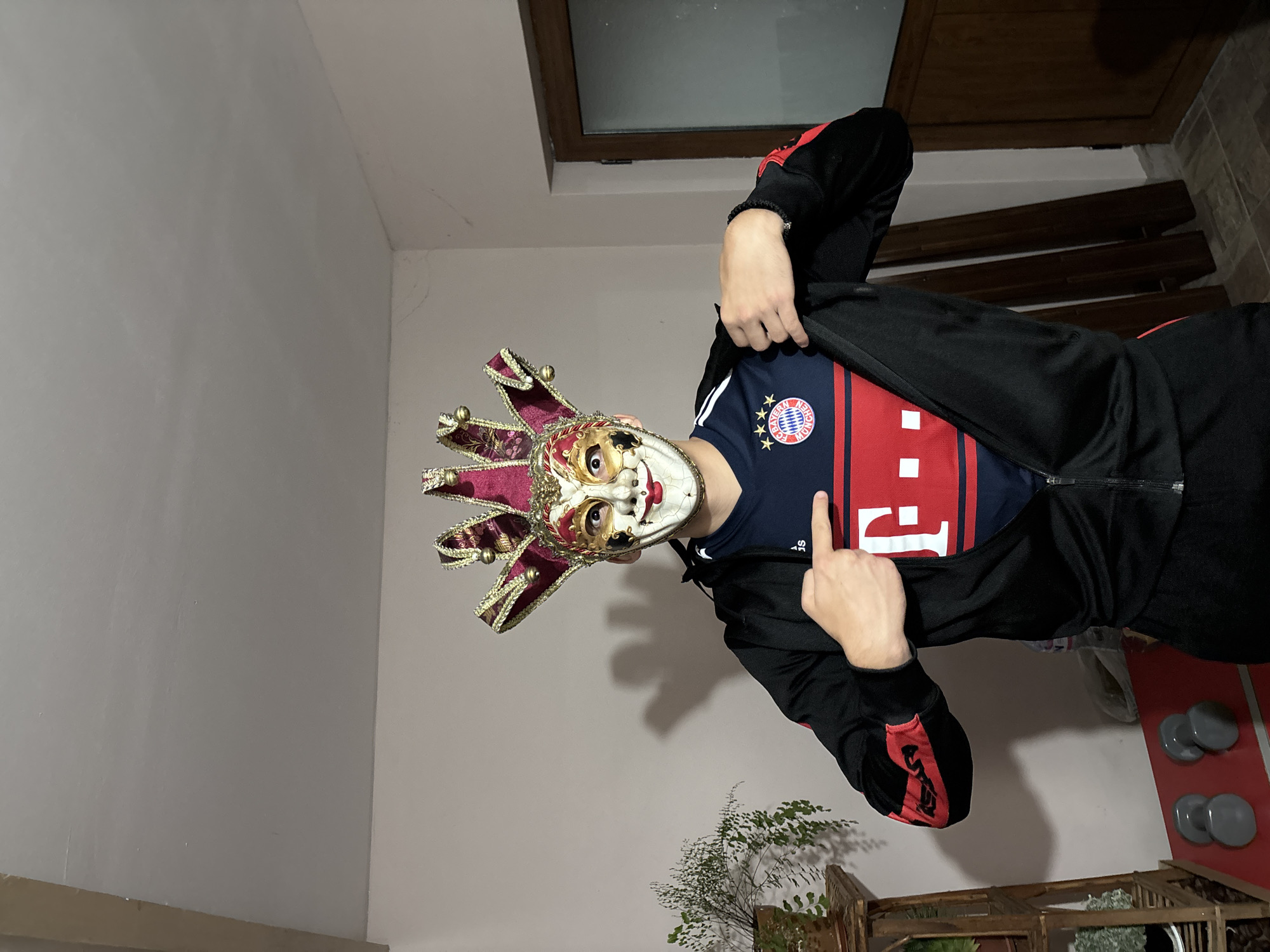 A person wearing a red and gold jester mask and a dark jacket with red stripes stands indoors. The person is pointing upwards with their right index finger. They are wearing a blue soccer jersey with a red stripe. In the background, there's a plant, a window, and a wooden structure. The specific location in Goritsa, Bulgaria is not discernible from the image.