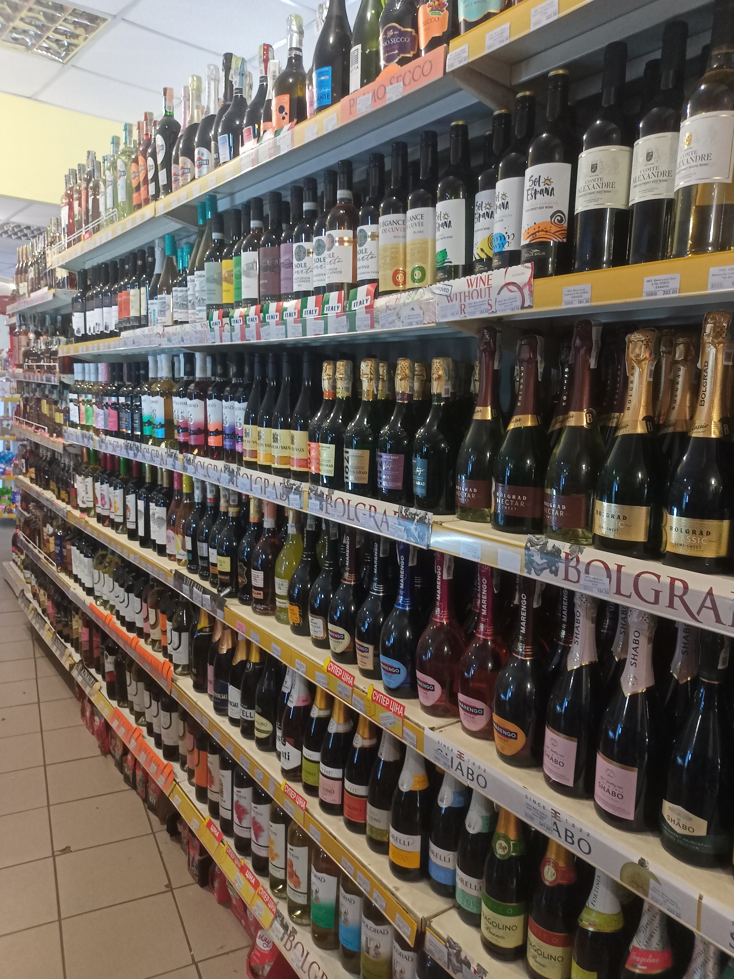 This is a medium shot of a well-stocked wine aisle in a supermarket in Bilhorod-Dnistrovskyi, Ukraine. The shelves are filled with numerous bottles of wine and sparkling wine, arranged by type and brand. The lighting is fluorescent, typical of an indoor retail environment, and the floor is tiled in a light beige color. The shelves are made of metal and are tiered, with yellow price tags visible on some of the lower shelves. The overall impression is one of abundance and variety, with a focus on alcoholic beverages. There are no people visible in the frame. The merchandise is predominantly in dark glass bottles, with some clear glass bottles also present. The labels on the bottles display a variety of languages, including Cyrillic and Latin scripts, indicating a diverse product offering.