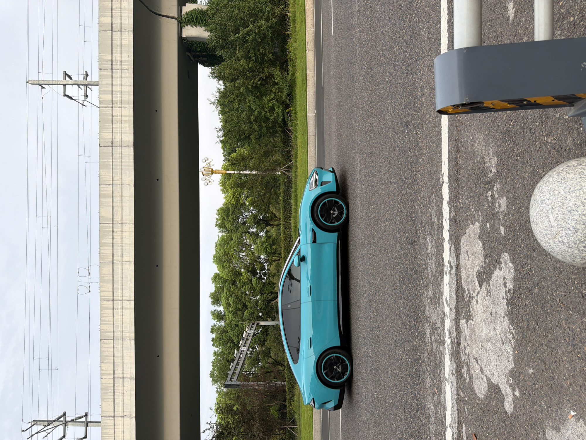 An electric blue car with black and blue wheels is driving on an asphalt road under an elevated highway or bridge. The car is captured in a side profile, moving from left to right. The background features lush green trees and vegetation, with power lines and a concrete structure of the overpass visible above. The sky is overcast. In the foreground, part of a road barrier and a large stone bollard are visible. This scene is located in Jiangjin District, Chongqing, China, as indicated by the distinct power line towers and infrastructure commonly found in the region.