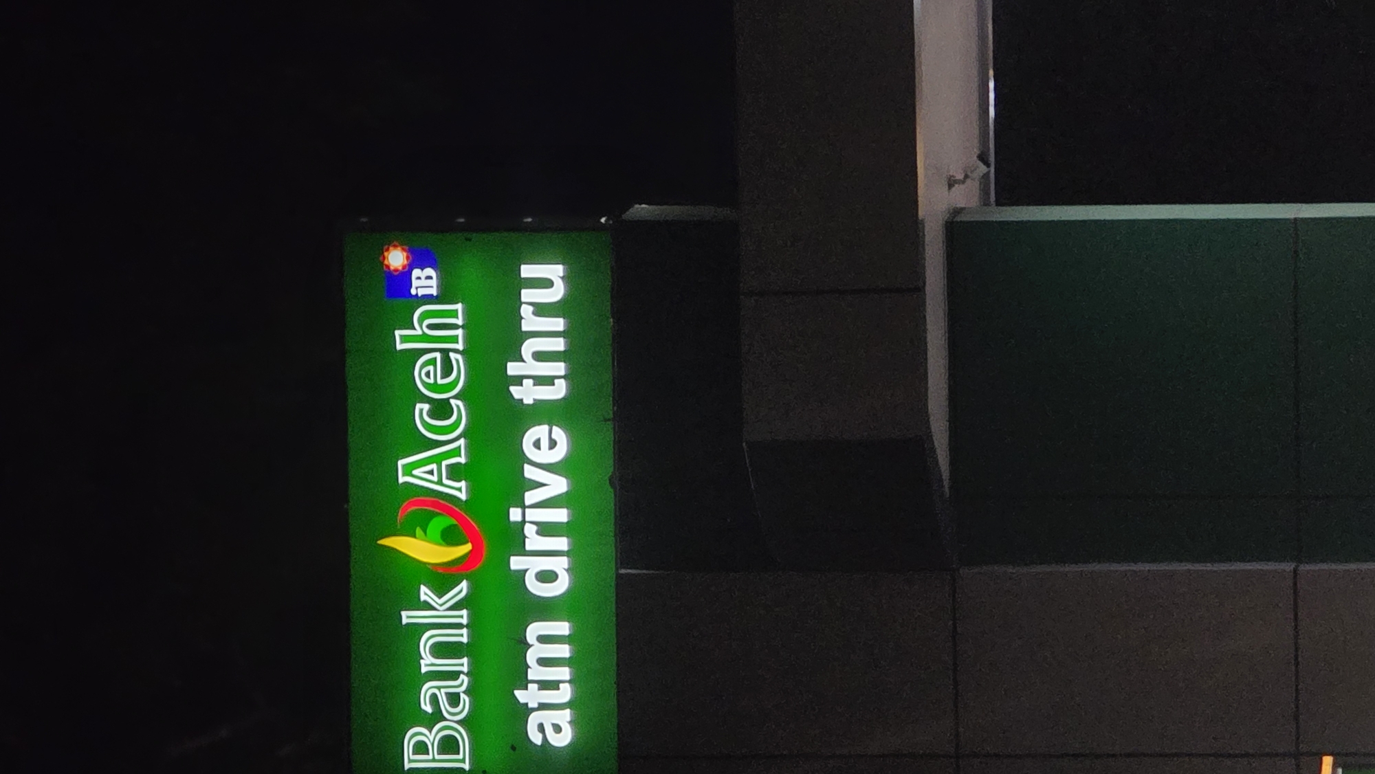 A nighttime shot depicts a Bank Aceh IB ATM drive-thru sign in Lhokseumawe, Indonesia. The sign is green and features the bank's logo, with the words "Bank Aceh IB" in green letters with a logo above, followed by "atm drive thru" beneath. The sign is illuminated. The environment is dark, with a tiled building to the right of the sign.