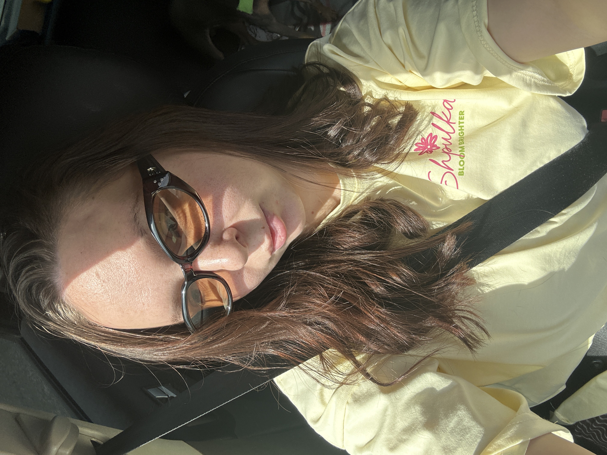 A person wearing a yellow t-shirt with the words "Shpulka Bloom Brighter" and sunglasses is lying down, secured by a black seatbelt. The environment appears to be the interior of a car. There is no information in the image to determine the location is Кельчино, Russia.