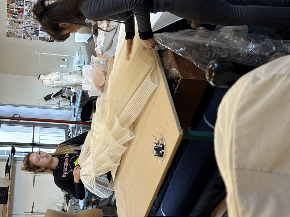 The image depicts an interior scene within a fashion or textile design workshop located in Arcueil, France. Two women are actively engaged in working on a large, off-white, intricately pleated fabric garment or textile piece laid flat on a light-colored wooden work table.
The woman on the left, of East Asian descent, has light brown hair tied in a bun and wears glasses. She is dressed in a black long-sleeve shirt with a yellow "PULCINO PI" graphic and is positioned behind the table, handling an upper section of the pleated fabric.
The woman on the right has dark hair tied up and is wearing a dark grey ribbed long-sleeve top and dark pants. She stands to the right of the table, using both hands to manipulate a lower section of the pleated fabric.
A black smartphone with white wired earbuds rests on the table surface, positioned between the two women and the fabric item.
In the background, behind the woman on the left, a window shows an indistinct building exterior. Nearby, a sewing machine and a mannequin or dress form are visible. Behind the woman on the right, another dress form marked with red lines, a thread stand, and other sewing equipment are present. The wall behind them is adorned with numerous pinned-up fabric swatches or design inspirations.
Underneath the work table, several rolls of dark blue fabric are stacked, and a wooden box containing various colored textile scraps or threads is partially visible. A clear plastic bag rests near the right woman's legs. The shoulder and upper back of a third individual, clad in a light-colored garment, are visible in the immediate foreground, out of focus.