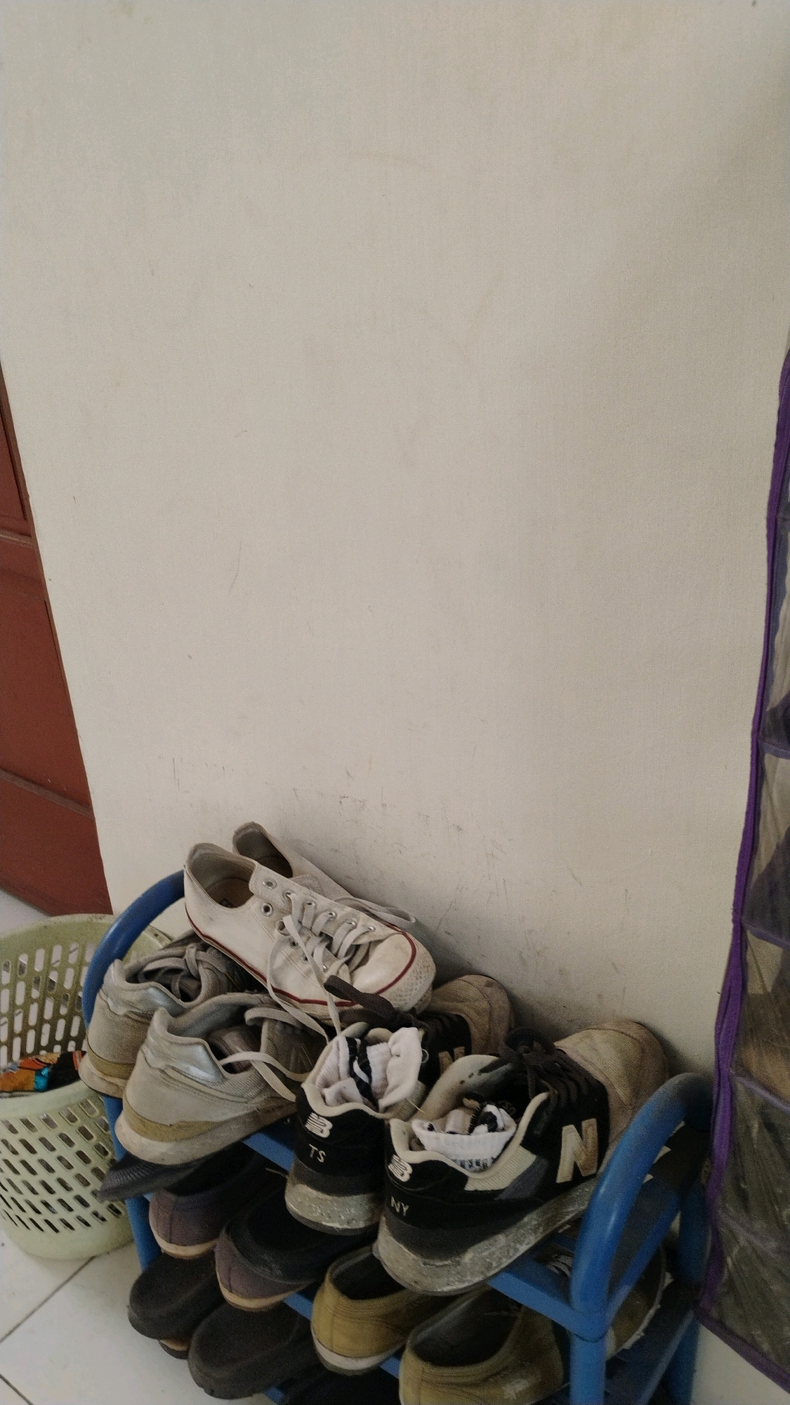 The image shows a blue shoe rack filled with various shoes and sneakers. On the top shelf, a pair of white Converse sneakers are prominently displayed, with their laces loosely tied. Below them, several other shoes are stacked, including what appear to be athletic shoes and casual footwear in various colors like beige, black, and gray. To the left of the shoe rack, a white plastic laundry basket is partially visible, containing some items. The shoe rack and basket are positioned against a plain, off-white wall. A brown door frame is visible on the far left edge of the image, and a section of a purple hanging shoe organizer is on the right side. The scene appears to be an indoor setting, likely a home or a common area, in Gumpang, Indonesia.