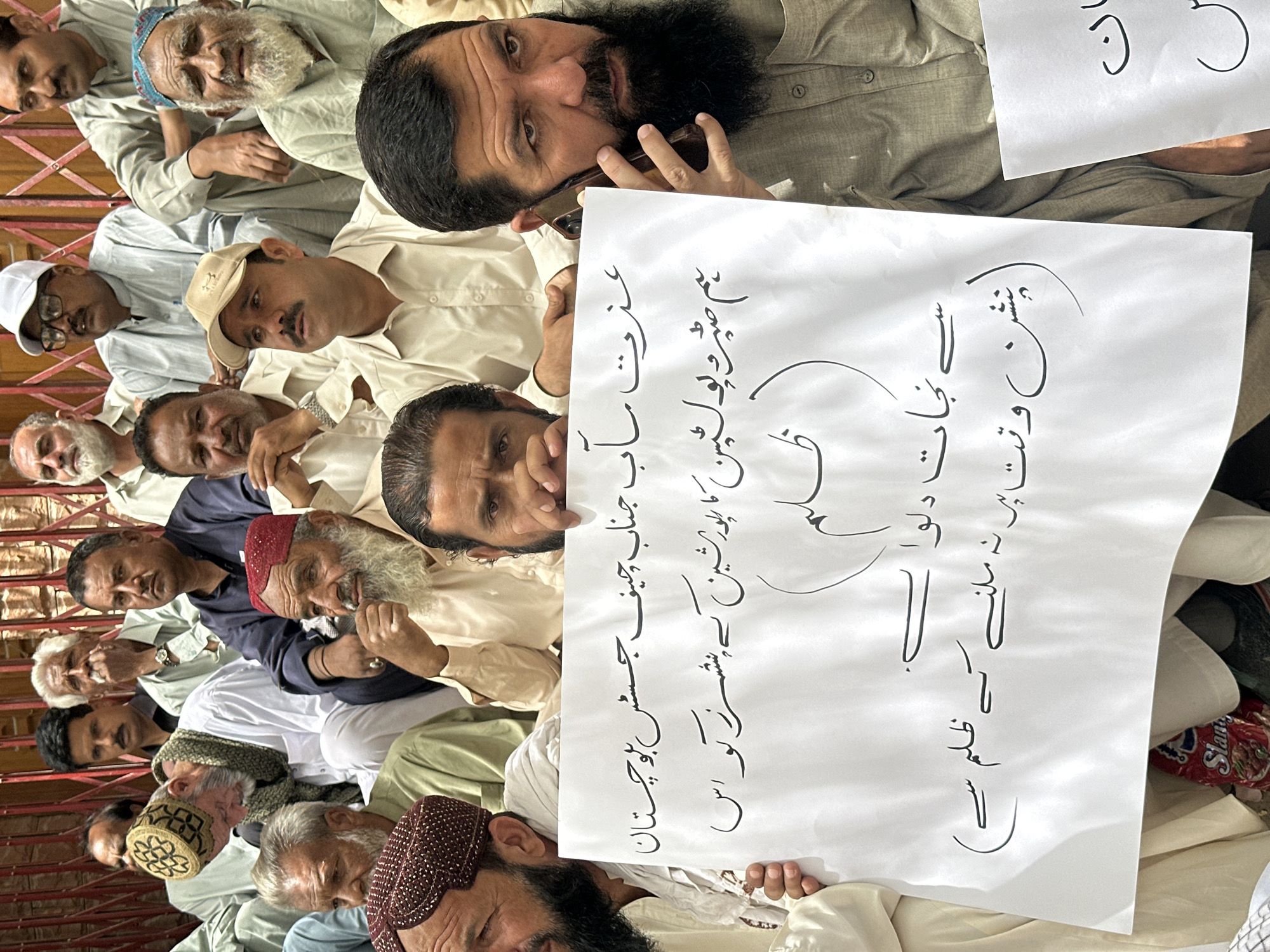 The image captures a group of men in Quetta City Tehsil, Pakistan, participating in what appears to be a protest. The men are primarily of middle age or older, with diverse facial features and attire, including traditional shalwar kameez and headwear. One man in the foreground is speaking on a mobile phone while holding a white placard. The placard has handwritten text in Urdu. Another man is partially visible holding a second placard. The men are seated or standing, and the background suggests an outdoor setting with a red lattice-like fence or wall. Some men are looking towards the camera, while others are engaged in their own actions. The overall atmosphere suggests a gathering expressing a grievance or demand.