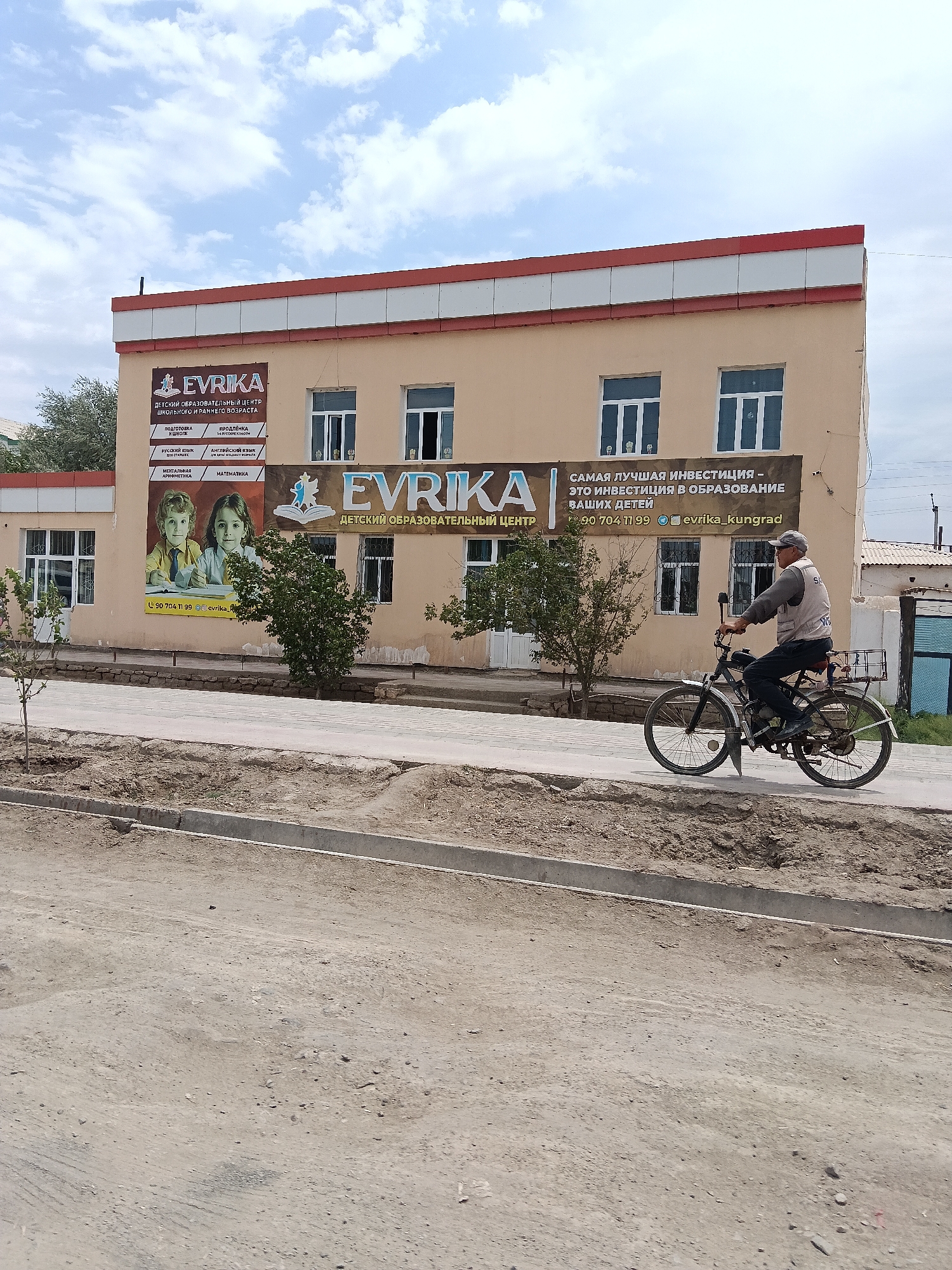 The image shows a street scene in Kungrad, Uzbekistan, featuring a two-story building with signage for a children's educational center. The building is painted beige with a red and white striped trim at the top. On the left side of the building, a banner displays "EVRIKA" along with text in Russian, advertising the "CHILDREN'S EDUCATIONAL CENTER OF SCHOOL AND EARLY AGE." Below this, there are smaller sections listing subjects such as "PREPARATION FOR SCHOOL," "RUSSIAN LANGUAGE," "MENTAL ARITHMETIC," "MATH," "ENGLISH LANGUAGE," and "SPEECH DEVELOPMENT." A large image of two smiling children is also part of this banner. Further to the right, another banner reads "EVRIKA" with the tagline "THE BEST INVESTMENT IS AN INVESTMENT IN THE EDUCATION OF YOUR CHILDREN," also in Russian, followed by a phone number and an Instagram handle "@evrika_kungrad."
In front of the building, a man wearing a cap and a grey vest over a long-sleeved shirt is riding a bicycle. The bicycle has a basket on the back and appears to be a moped or motorized bicycle. He is traveling from left to right across the frame.
The environment includes a paved sidewalk in front of the building, with a dirt area and a curb bordering it. The street in the foreground is unpaved and appears dusty. Trees are visible along the sidewalk. The sky is partly cloudy. The overall impression is an urban street on a bright day, with evidence of ongoing development or maintenance in the foreground.