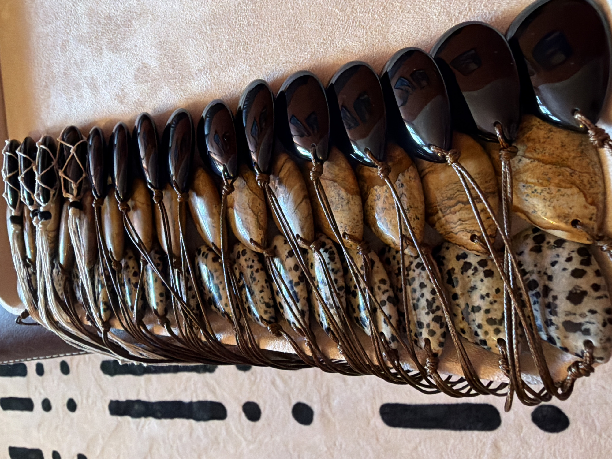 The image displays a close-up, overhead view of a collection of pendants or beads strung together on thin cords. The pendants are a mix of two types: some are smooth, teardrop-shaped, and dark brown or black, possibly obsidian or a similar polished stone. Others are oval-shaped and appear to be made of a mottled, earthy-toned stone with brown and black speckles, resembling jasper or a similar patterned agate. The pendants are arranged in a curved line, suggesting they might be part of a necklace, bracelet, or a decorative display. The cords are a neutral brown, tied in a way that secures each pendant individually. The background is a textured, light-colored surface, possibly fabric or a natural material, with a darker, patterned area visible at the bottom edge of the frame, featuring black rectangular shapes and dots. The lighting suggests an indoor setting, with reflections visible on the polished dark pendants. No people are visible, and the focus is entirely on the arrangement of the pendants and their texture. The scene is static, with no activity taking place. There is no visible text in the image. The context provided suggests the location is Khimki, Russia.