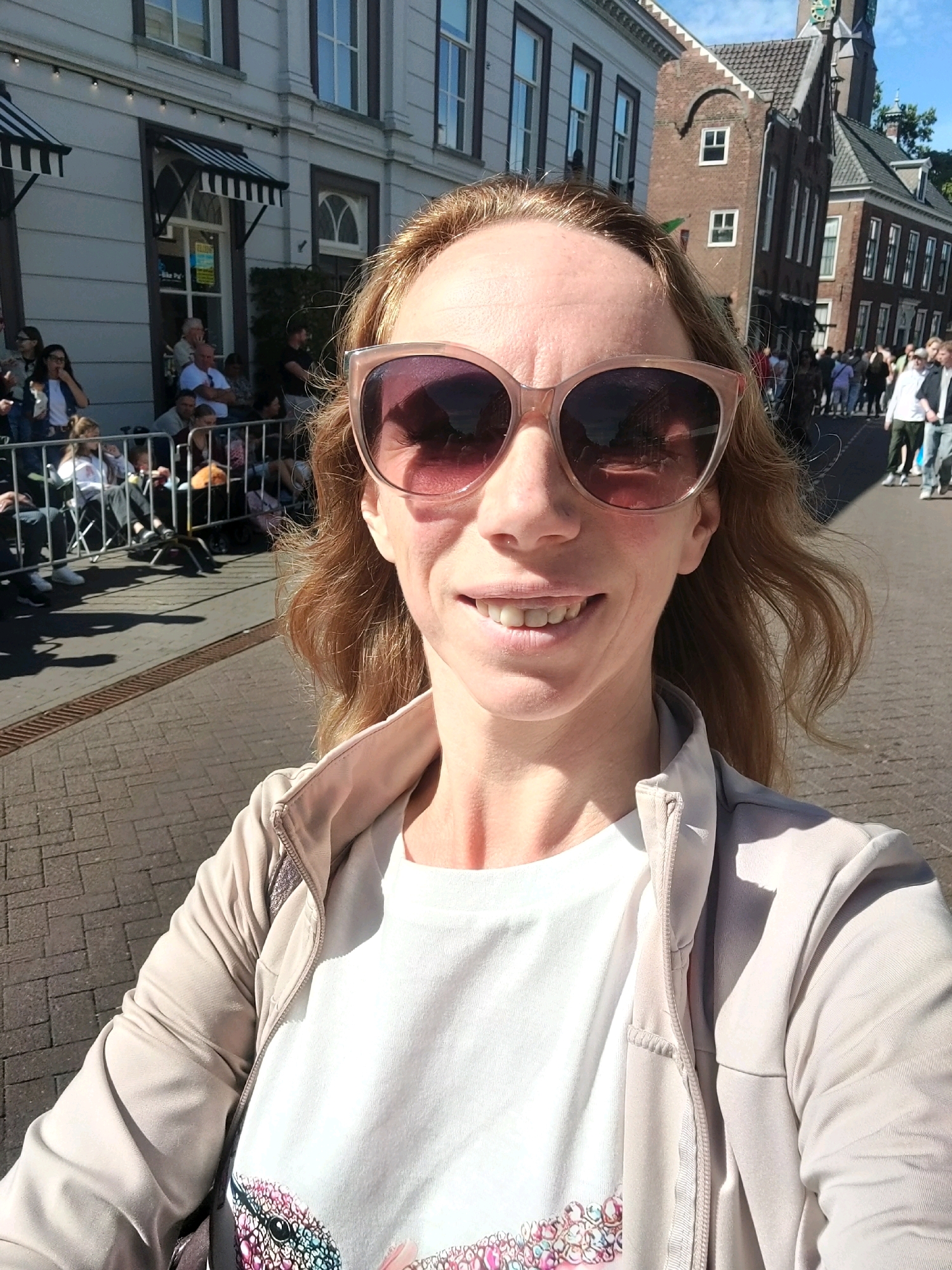A woman with shoulder-length wavy blonde hair, wearing oversized pink-tinted sunglasses and a light beige jacket over a white t-shirt with a colorful hummingbird graphic, takes a selfie outdoors. She is smiling broadly, revealing her teeth, and her face is illuminated by sunlight. In the background, a crowd of people is visible, some standing and others seated behind a metal barrier, suggesting a public event or parade. The setting appears to be a street in a European town, characterized by brick pavement, buildings with windows and awnings, and a clock tower in the distance. This scene is consistent with the annual Flower Parade in Zundert, Netherlands, where spectators gather along the route.
