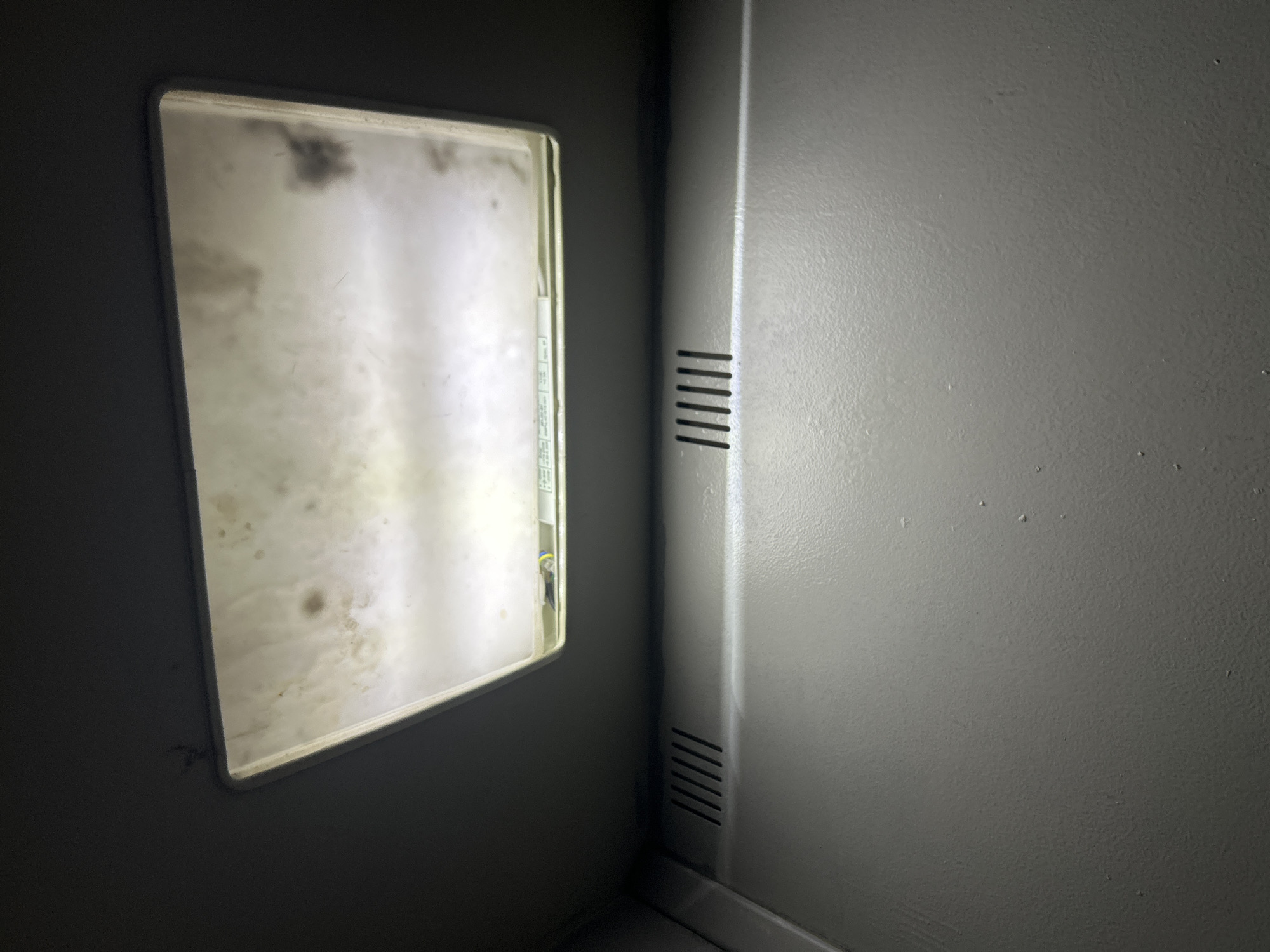 The image shows an interior corner of a room, possibly a utility or service area. The walls are painted a dull gray. On the left is a rectangular light fixture recessed into the wall, its diffuser heavily soiled with grime and dark spots. The fixture emits a pale, diffused light. Adjacent to the fixture on the right is a vertical section of the wall with horizontal ventilation slots near the top and bottom. The lighting is dim and uneven, casting shadows and highlighting the texture of the painted surfaces. The overall impression is one of neglect and poor maintenance. There is no evidence to determine the location is in Moscow, Russia.
