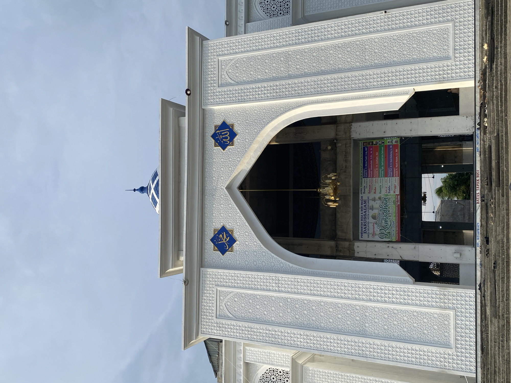 The image shows the exterior of a mosque in Lhokseumawe, Indonesia, featuring a white facade with intricate geometric carvings and Islamic calligraphy. A blue-tiled dome is visible at the top. An arched entrance leads into the mosque, with a chandelier hanging inside. A banner with the text "Ramadhan" is displayed near the entrance. The steps leading up to the mosque are visible at the bottom of the frame, with several pairs of sandals placed on them. The sky is overcast.