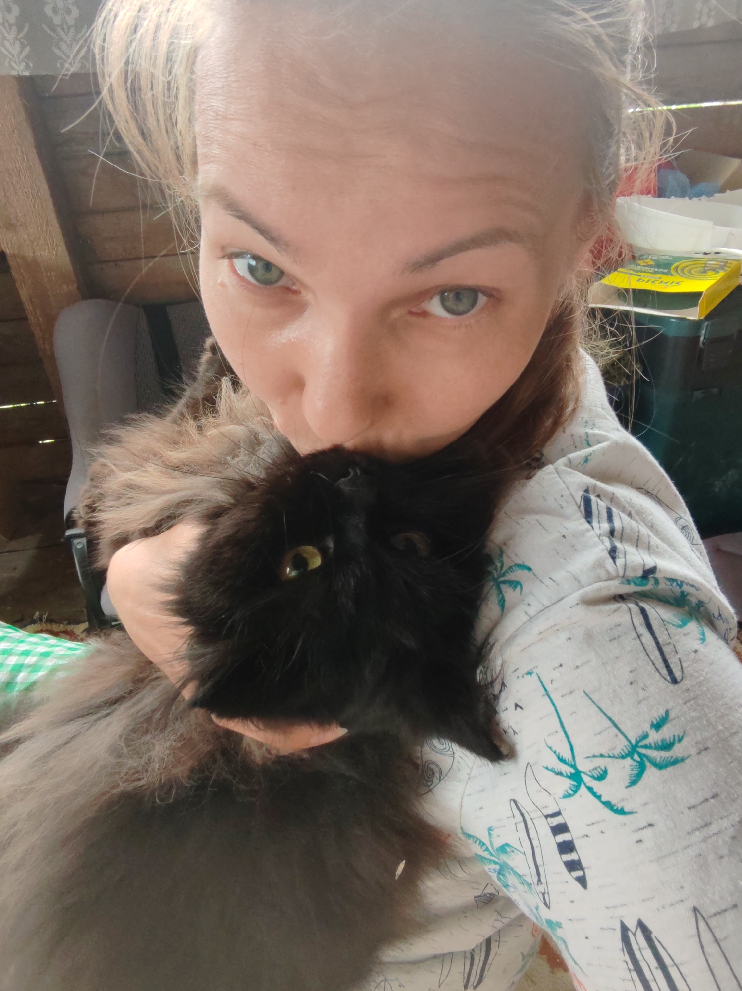 A close-up, selfie-style portrait shows a person holding a black cat. The person's face is partially obscured by the cat, but their green eyes are visible, looking directly at the camera. The person's blonde hair is pulled back loosely. They are wearing a light-colored shirt with a pattern of palm trees and surfboards. The cat is a fluffy, dark-colored breed with yellow-green eyes. The background is blurred and appears to be an indoor setting with wooden walls and shelves, possibly in a rural or older building. A green and white checkered cloth is visible in the lower left corner. The overall impression is of a casual, affectionate moment between a person and their pet. The context of "Чухлэм, Russia" suggests this scene is taking place in a residential or domestic environment within that specific location.