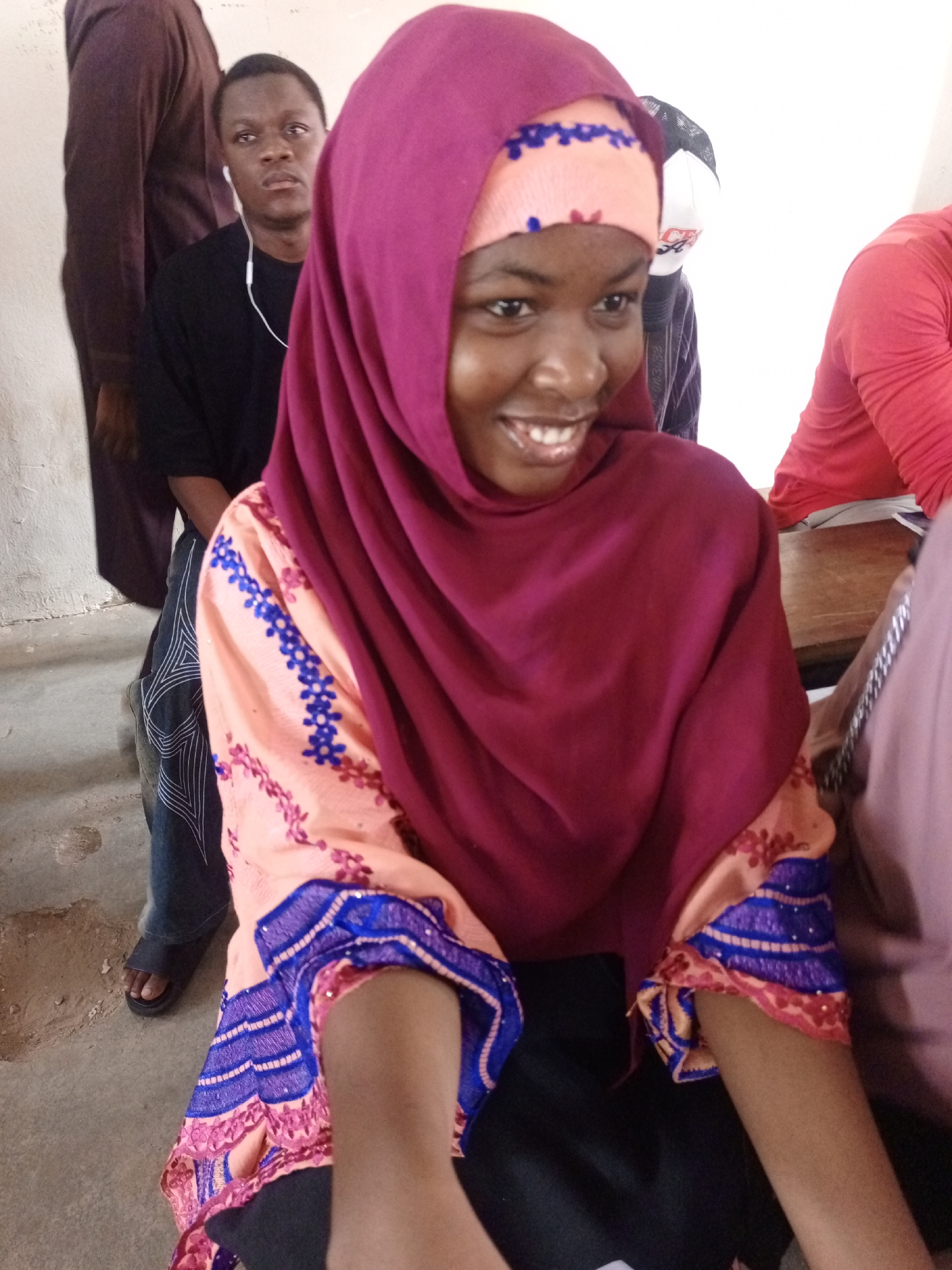 This indoor photograph, taken in Keffi, Nigeria, features several individuals in what appears to be a communal gathering space. In the foreground, a young Black woman is smiling broadly, looking to her right. She wears a deep maroon hijab over a light orange patterned headscarf and a long-sleeved light peach-colored top adorned with blue and pink/red embroidery. Black trousers are visible beneath her top.
To her left, a young Black man sits, wearing a black t-shirt and white earbuds. He looks forward with a neutral to slightly serious expression. His dark jeans, featuring distinctive white stitching patterns, and black sandals are visible. Behind him, a person in a dark brown traditional garment is partially seen. Further in the background, another individual wears a white baseball cap with a dark brim and partially obscured text, along with a dark top. On the far right, segments of other people in red and light purple attire are visible, along with a wooden surface, likely a table or desk.
The setting includes light-colored walls and a concrete floor exhibiting some wear. The overall atmosphere suggests a casual gathering, perhaps a class or social meeting, marked by the prominent cheerful expression of the woman in the foreground. The attire of the subjects is consistent with the cultural context of Nigeria. There is no clearly readable text in the image.