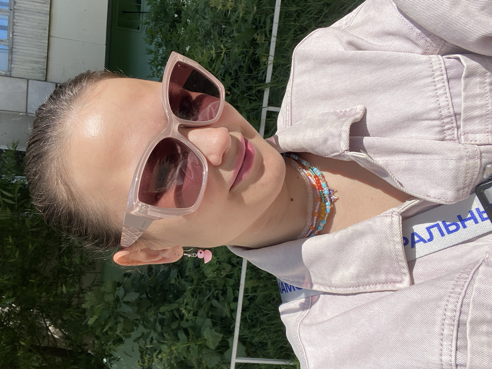 This is a portrait of a young woman, taken from a close-up, low-angle perspective. She is wearing large, light pink, cat-eye sunglasses and a light purple denim jacket. Her hair is pulled back, and she has a pink flamingo charm earring in her left ear. She is also wearing a beaded bracelet with colorful beads and charms. Her lips are slightly parted, and she appears to be looking towards the camera. The background consists of greenery, suggesting an outdoor setting, possibly in a park or garden. Sunlight is casting shadows, indicating it is daytime, likely during sunny weather. A white metal fence is visible in the background. A blue tag with white Russian text that reads "РАЛЬНЕ" is attached to the jacket. The overall impression is casual and summery, with a playful, youthful aesthetic. The location context is Chaikovsky, Russia.