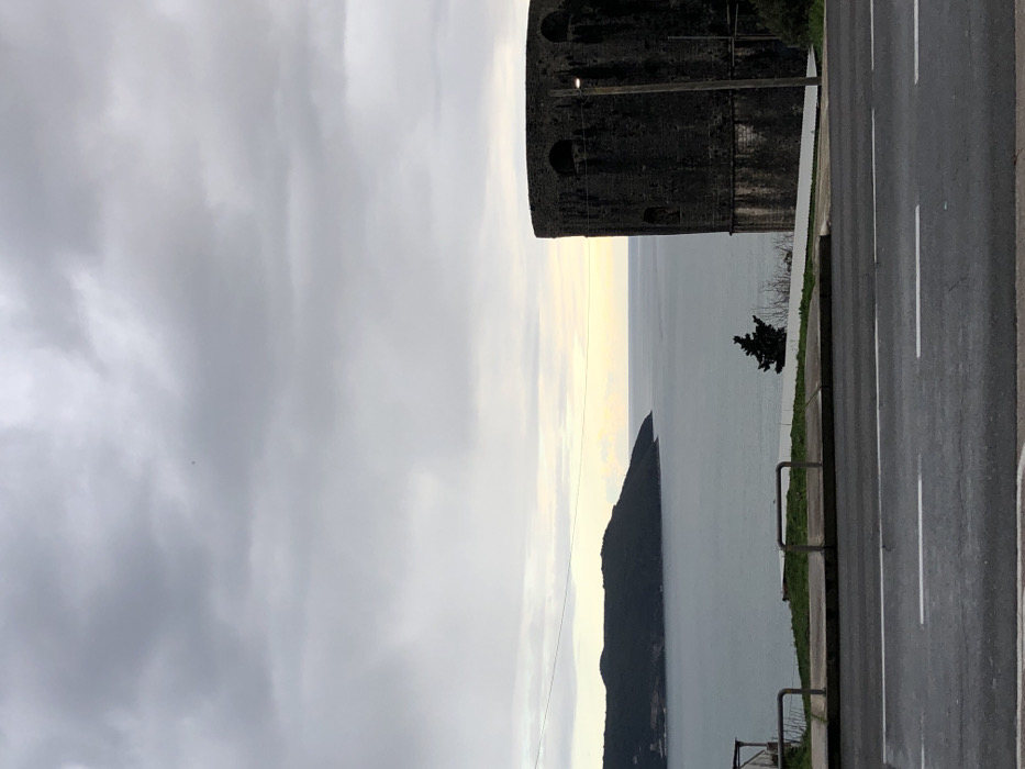 A roadside view features a stone tower near a body of water. An island, shaped like a jagged peak, stands in the water. A road with white stripes runs along the edge of the frame. A few trees grow by the water’s edge. The sky is overcast with grey clouds.
