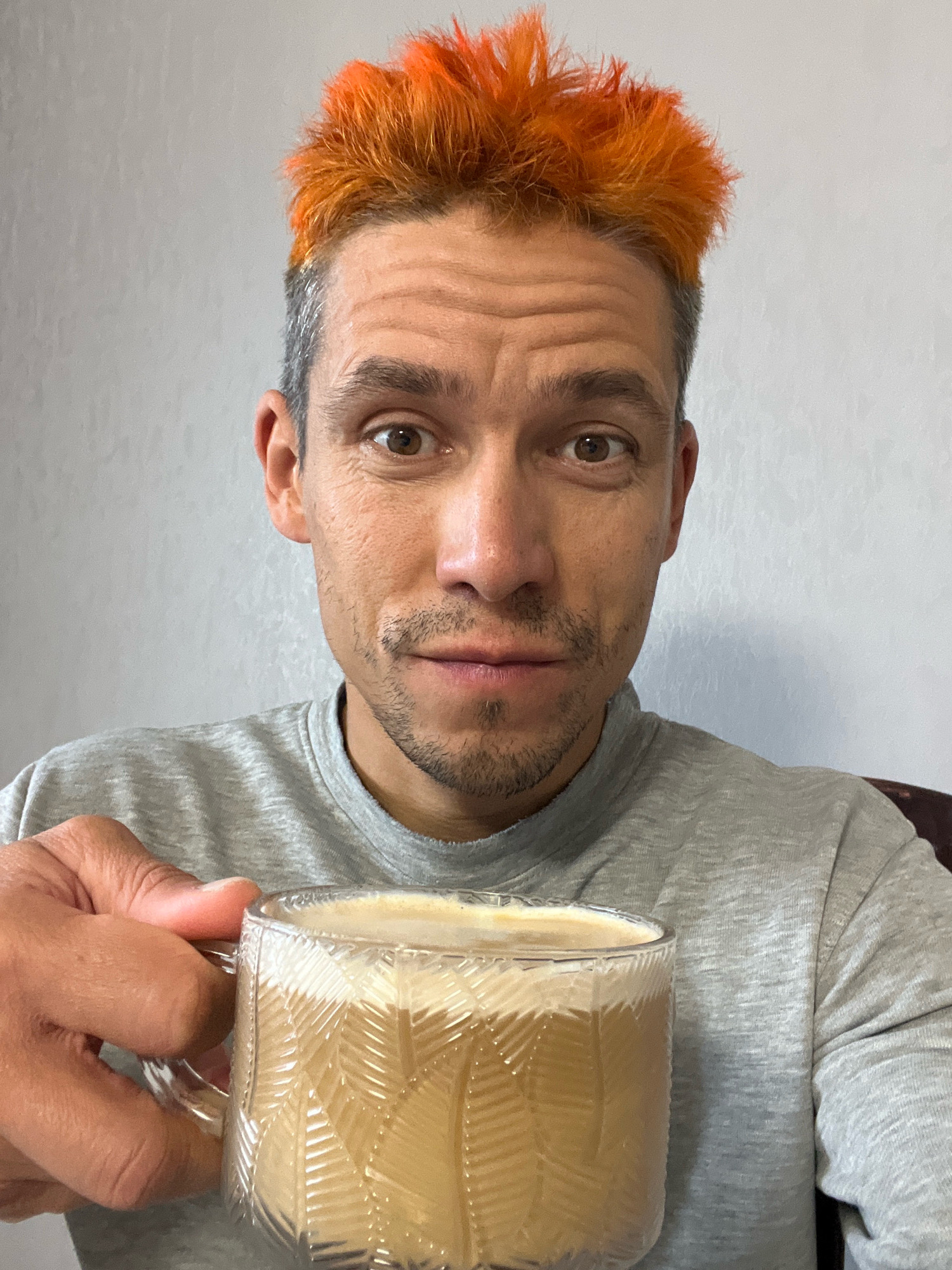 A man with brightly dyed orange hair and a grey shirt is holding a clear glass mug filled with a light brown, frothy beverage. The man is looking directly at the camera with a neutral expression. The background is a plain, textured grey wall. No specific location in Chaikovsky, Russia can be determined from the image.