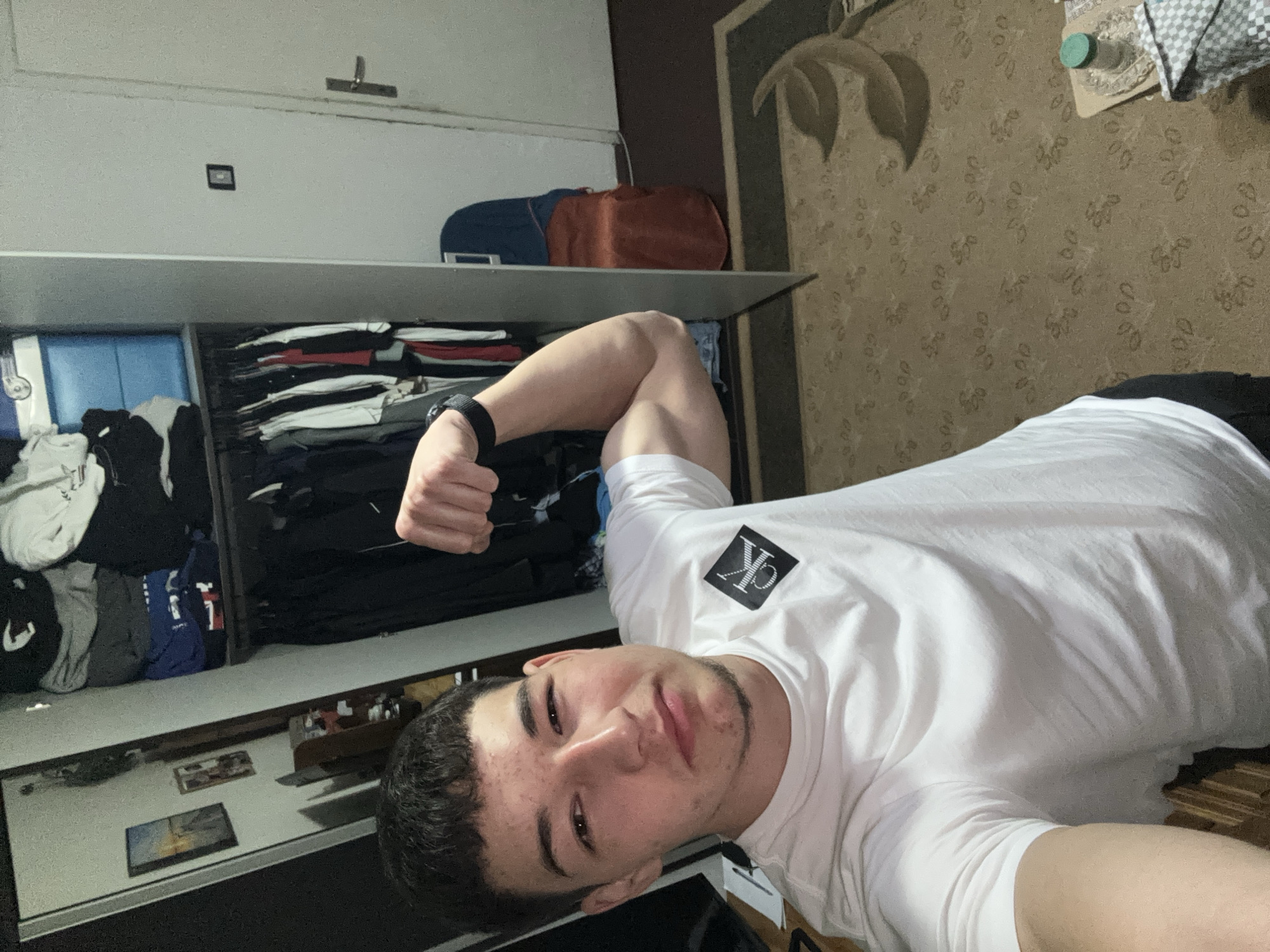 A young man wearing a white Calvin Klein T-shirt poses for a selfie indoors, flexing his bicep. A shelf system behind him holds clothing, bags, and decorative items. The room has patterned wallpaper and appears to be in a residence. The city of Zlatograd, Bulgaria is not discernible from the image.
