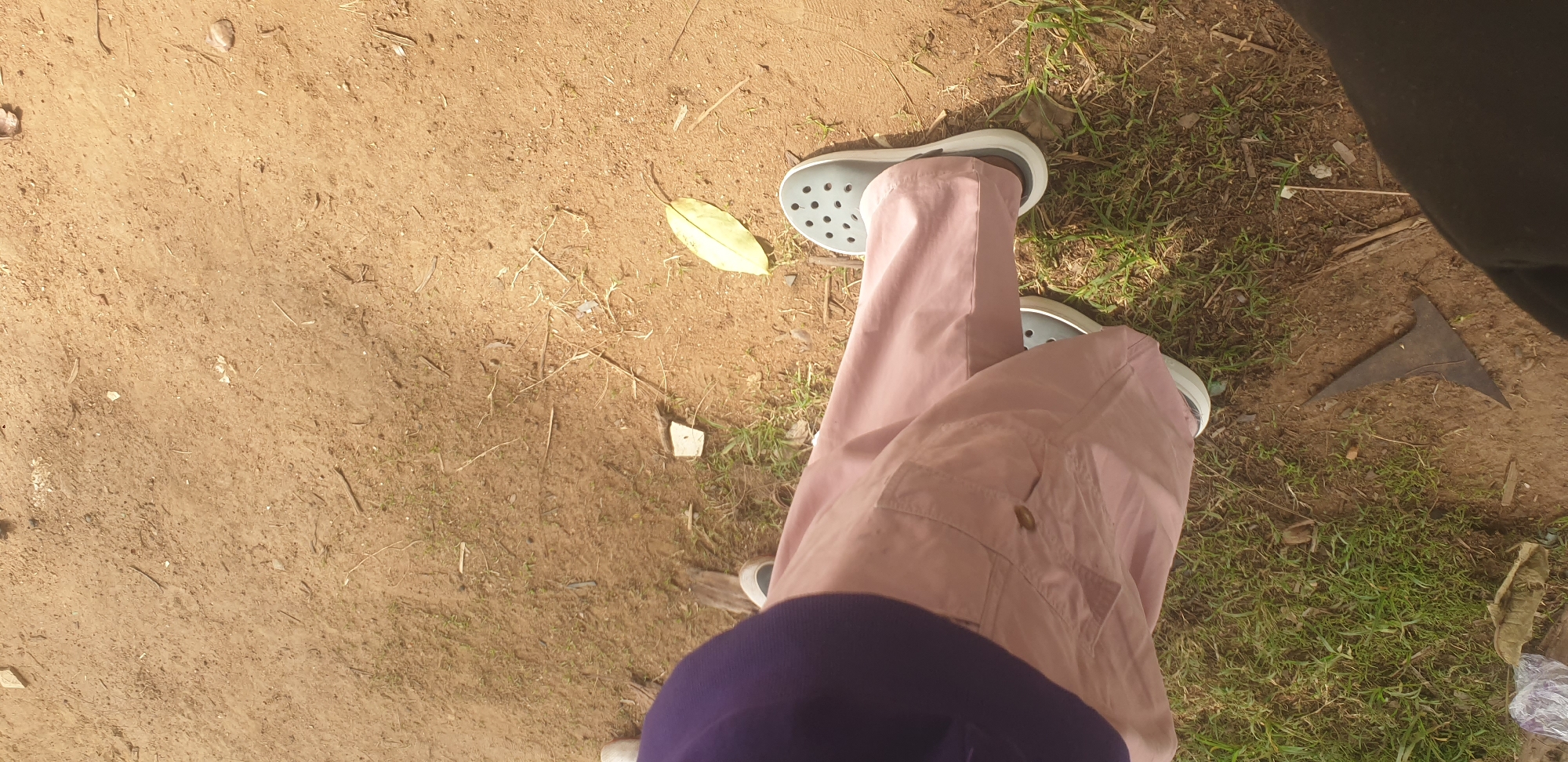 This media file depicts a person's legs and feet, shot from a top-down perspective. The individual is wearing light pink trousers and grey Croc-style sandals. They are standing on a dirt path with patches of green grass and dry leaves scattered around. To the right of the frame, a dark, triangular object made of metal lies on the ground. The scene appears to be outdoors, possibly in a rural or natural setting. The lighting suggests it is daytime, with sunlight casting shadows. No text is visible in the image. The overall impression is of a casual, everyday moment. The location context of Yelwa, Nigeria, further grounds the image in a specific geographical area.