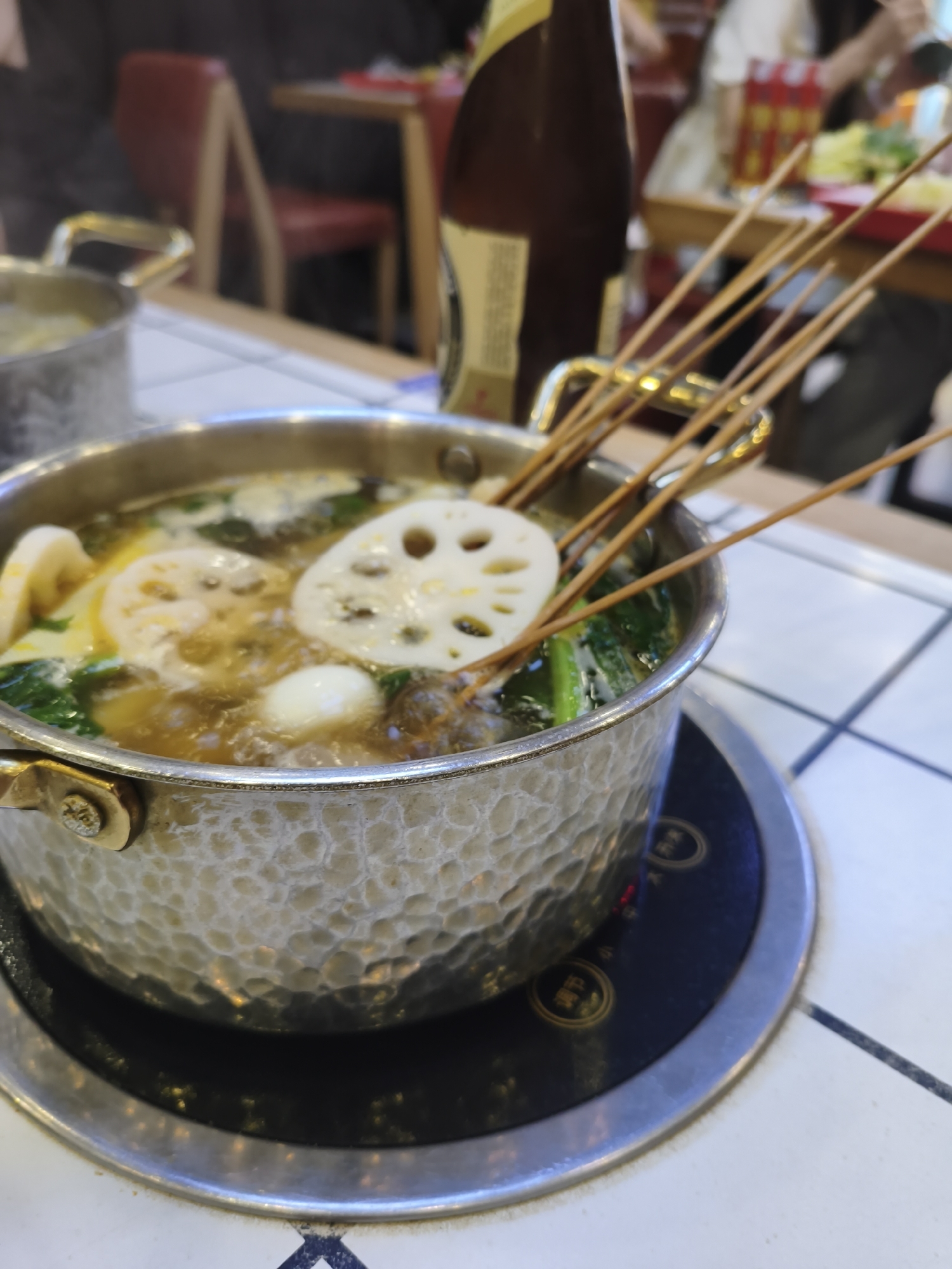 A close-up shot captures a steaming metal pot of hot pot, filled with broth, lotus root slices, quail eggs, and green vegetables, with wooden skewers emerging from the broth. The pot rests on a black electric burner, which is placed on a white tiled table. In the background, a dark brown bottle of liquid stands to the right of the pot. Blurred figures and a red chair are visible in the background, suggesting a restaurant setting. The overall scene depicts a meal being prepared or served, likely in Mudanjiang, China.