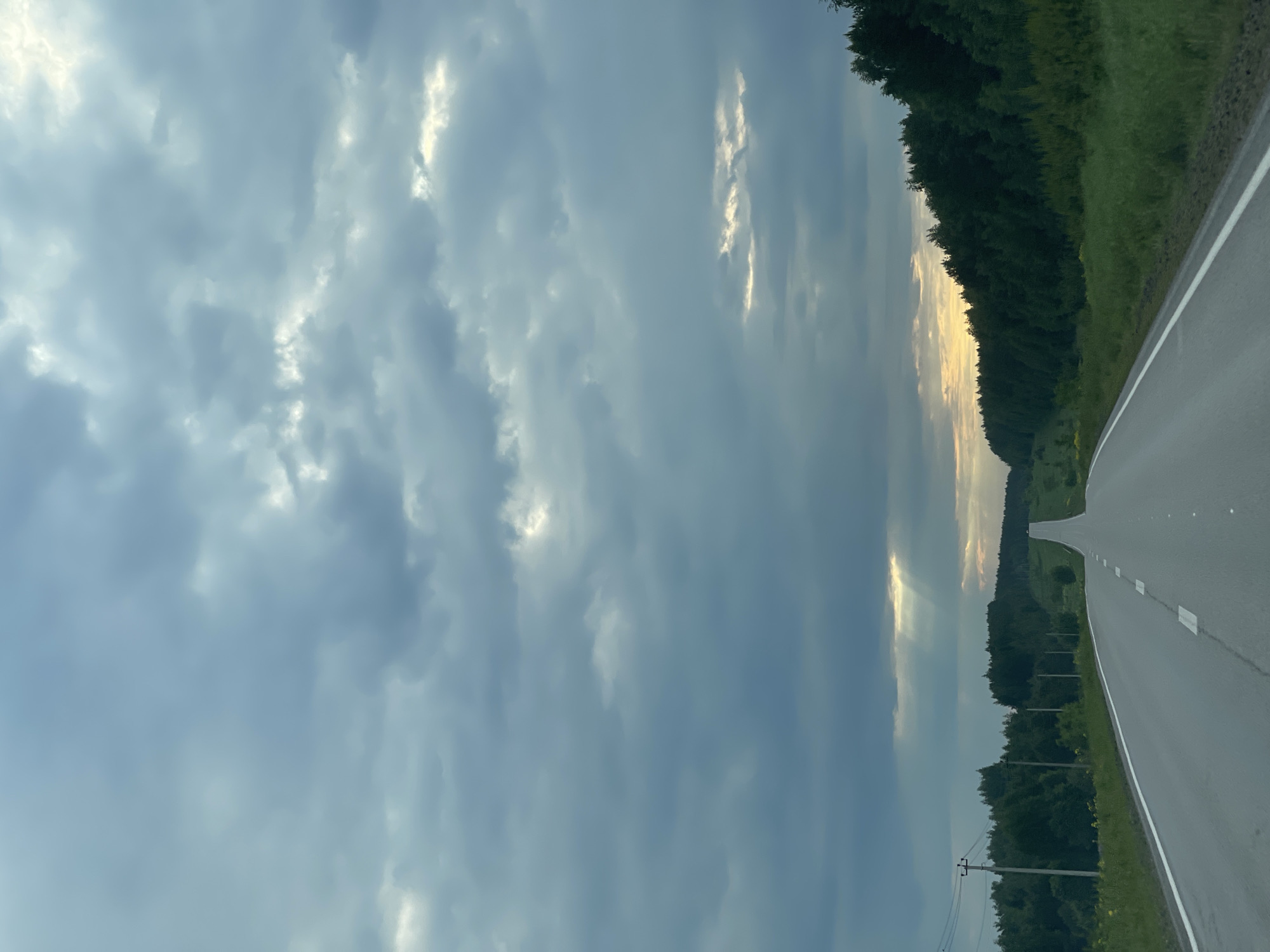 The image displays a scenic landscape featuring a paved road extending into the distance under a dramatic, cloudy sky. The perspective is from a vehicle, looking ahead as the road curves gently. White lane markings, including a solid line on the right edge and dashed lines further along the asphalt, are visible.
To the right of the road, a dense, dark green forest lines the horizon, with patches of lighter green grass and vegetation closer to the road. The sky is predominantly covered by heavy grey and blue clouds, indicating an overcast day. However, rays of golden light pierce through the clouds in the distance, illuminating parts of the sky and suggesting the time of day is late afternoon or early evening, possibly approaching sunset. On the left side of the road, electrical utility poles with wires are visible, hinting at a rural or semi-rural setting.
No people or discernible text are present in the image. The scene conveys a sense of peaceful journey through a natural, undeveloped environment, with the dynamic sky adding to its atmospheric quality.