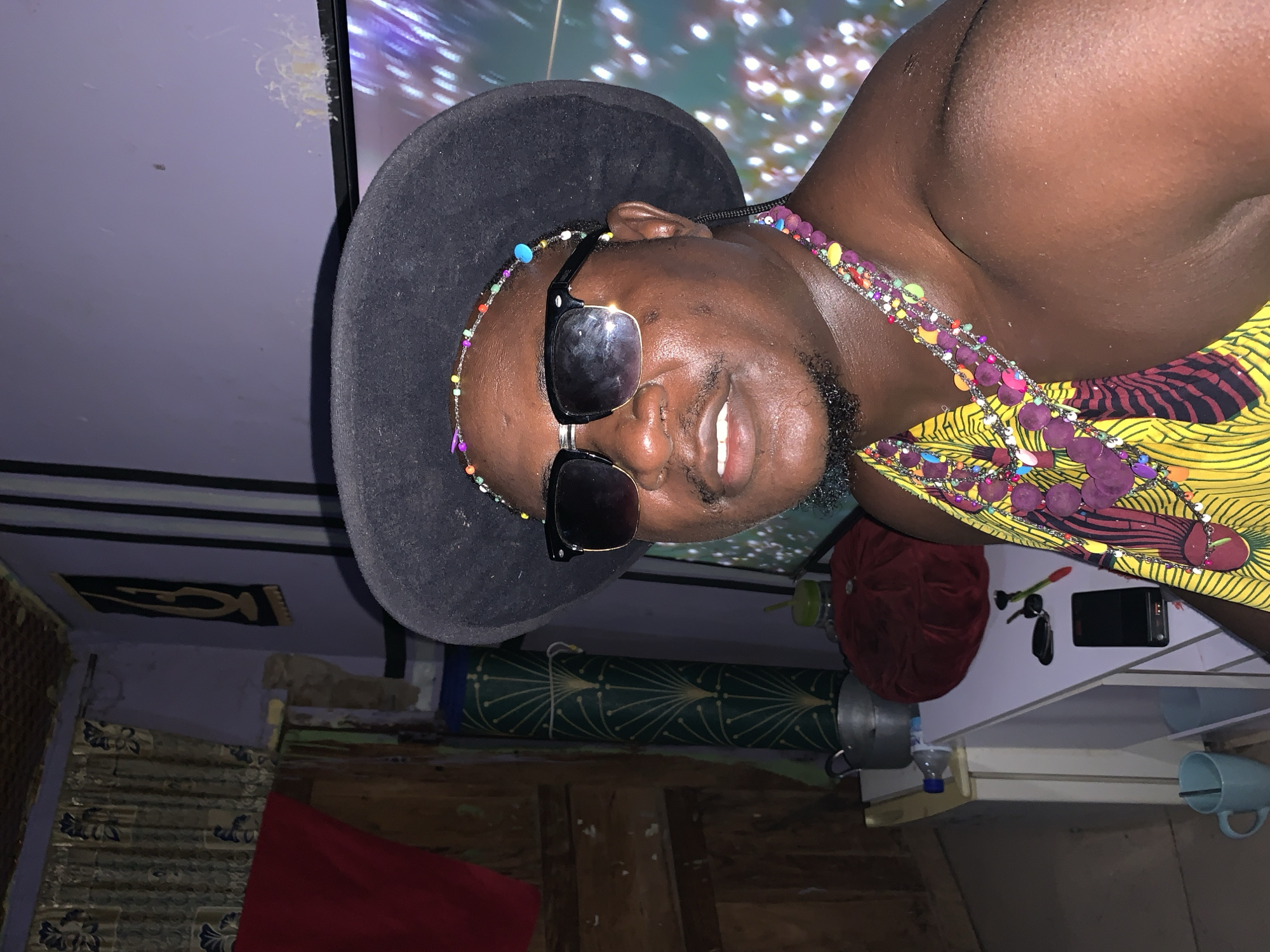 A medium shot shows a dark-skinned man with a goatee, wearing a black fedora and sunglasses, looking at the camera. He has a colorful beaded headband and a layered beaded necklace over his bare shoulders. He is wearing a yellow garment with a vibrant abstract pattern. He is indoors, possibly in a home in Akwanga, Nigeria. In the background, a television screen displays a blurry, colorful abstract image. To the left of the man, there is a wall with curtains and a red cloth hanging. Below the television, a white shelf unit holds various items, including a red cushion, keys, a small black electronic device, a plastic bottle, a metal pot, and a green straw in a bottle. A blue mug with a spoon is on the shelf below.