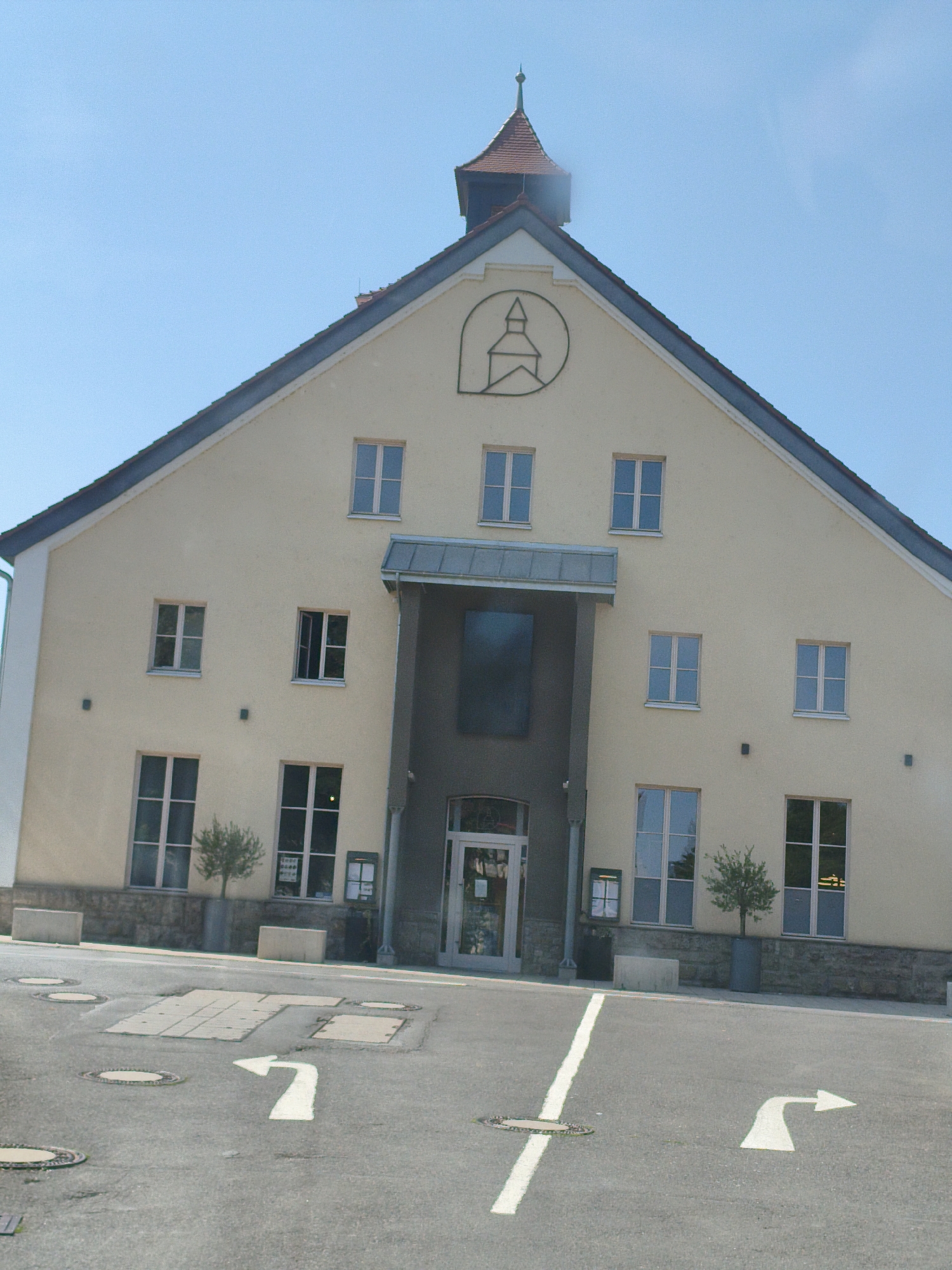The image shows a light yellow, two-story building with a dark roof and a pointed spire on top. The building has several windows and an entrance with a covered overhang. A symbol depicting a church tower is visible on the front of the building. In front of the building, there is a paved area with white directional arrows painted on the ground. Several circular manhole covers are also present. The sky is clear and blue. The building is located in the city of Ochsenfurt, Germany.