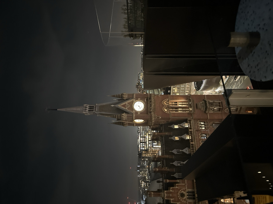 The photo showcases a building at night, possibly a historic clock tower, with prominent clock faces illuminated against the dark sky. The tower's architecture is detailed, with multiple tiers and spires. Contemporary buildings are visible nearby with lighted windows, hinting at an urban setting. A portion of a modern structure is in the foreground, including a glass panel and a circular table, suggesting the photo was taken from a rooftop bar or restaurant.