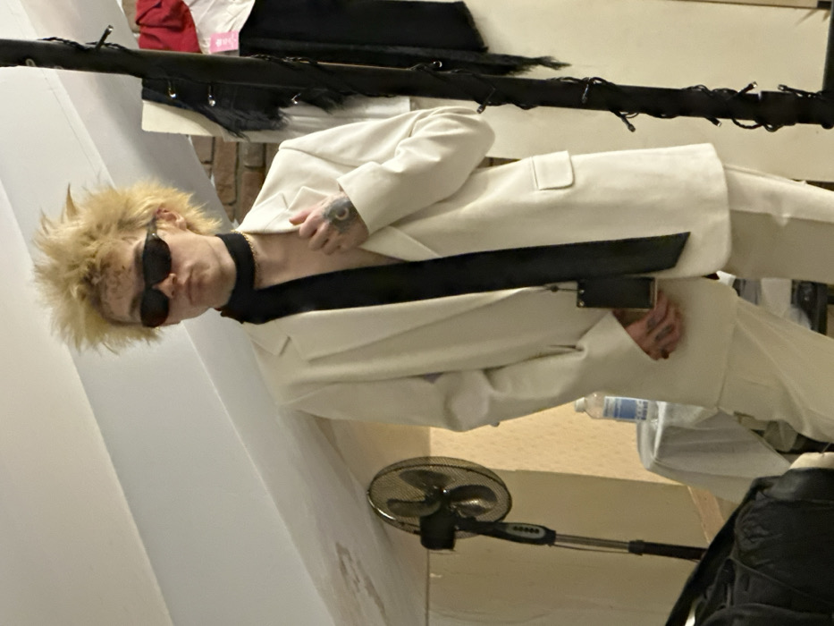 A person with spiky, blond hair and dark sunglasses poses in a white suit with a black top. The suit jacket is partially open, revealing a glimpse of their chest and a black choker. Tattoos are visible on their hand. The photo is taken indoors with clothing rack behind them. A standing fan is visible on the right side of the image. A water bottle is also visible in the background.