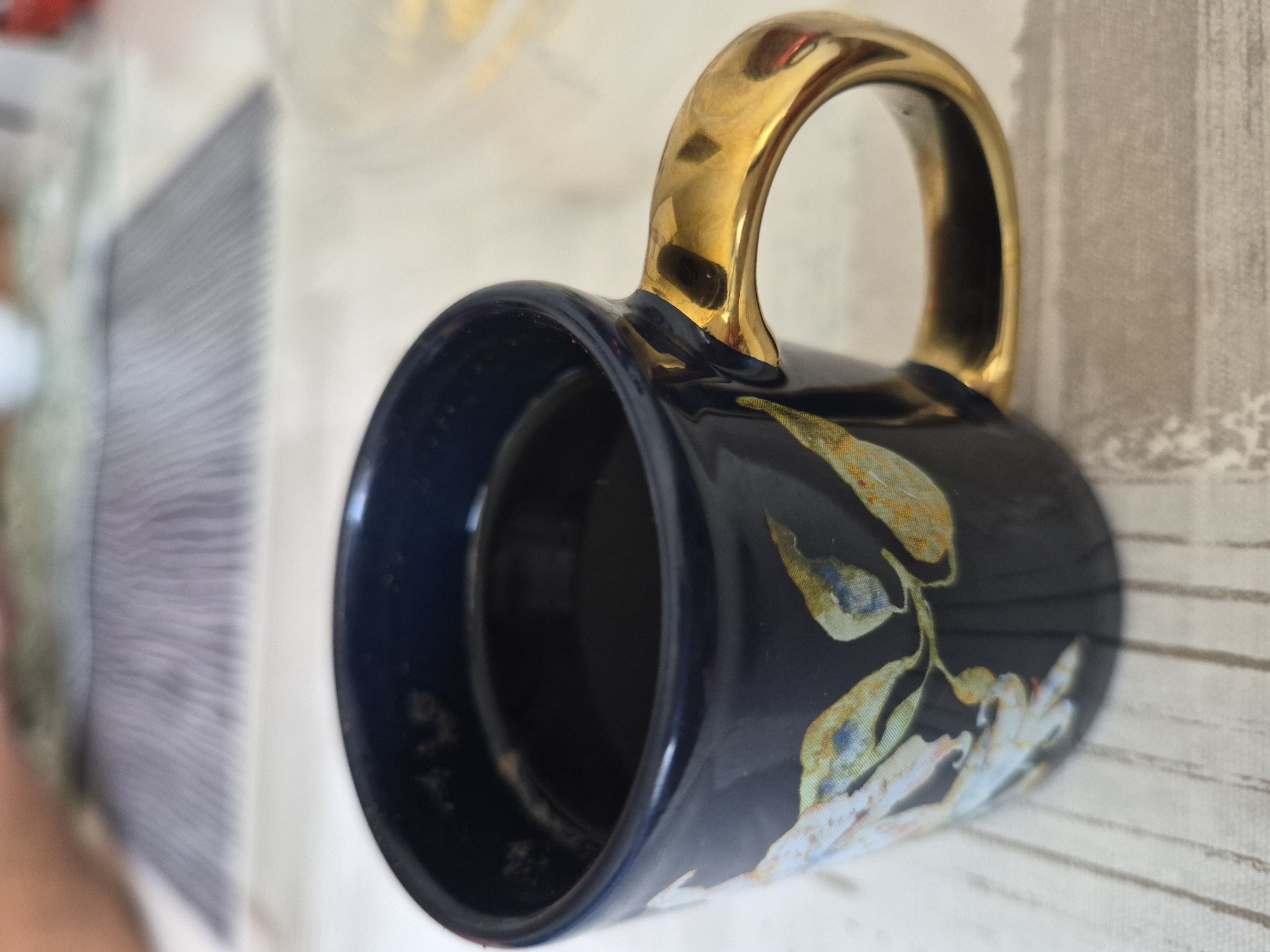 The media file shows a close-up, rotated view of a dark blue mug with a golden handle. The mug is decorated with a floral pattern featuring light-colored leaves with hints of yellow and blue. The background is blurred, but appears to be a textured surface, possibly a tablecloth, with subtle vertical lines. Another object, possibly another mug or a container, is visible and out of focus in the upper left of the image. The lighting suggests an indoor setting, possibly during the daytime. There are no people or discernible activities taking place. The focus is entirely on the mug and its decorative details.