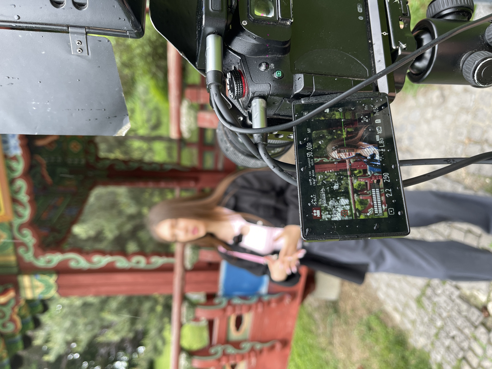 The image displays an outdoor video recording setup in Kyiv, Ukraine, captured from an overhead perspective and rotated 90 degrees clockwise. In the foreground, a professional mirrorless camera is visible, equipped with a matte box or lens hood. Several cables are connected to the camera body, and small water droplets are present on its surface, suggesting rain or recent rainfall.
The camera's articulating screen, also dotted with water, shows a live feed of the scene being filmed. Visible on the screen are various recording parameters: "50.00p" (frame rate), "MF" (Manual Focus), "C4K LongGOP" (video resolution and compression), "LPCM 150M" (audio bitrate), and "420/8" (color sampling). Other displayed information includes "1m25s" (recording duration), "L.8" (likely f/1.8 aperture), and "2.2 250" (possibly aperture and shutter speed). "NAT PEAK H", "Fn", "S", and "WIFI" buttons or indicators are also visible.
The subject of the recording, a young woman with long brown hair, is in the blurred background. She is wearing a dark jacket over a light top and appears to be holding a blue object, possibly a book or folder. The setting is an ornate traditional structure, possibly a gazebo or pavilion, characterized by red beams and intricate green and yellow decorative patterns. Beyond this structure, lush green foliage and trees are visible, along with stone paving on the ground. The image captures a professional video production in a scenic, possibly historical, outdoor location.