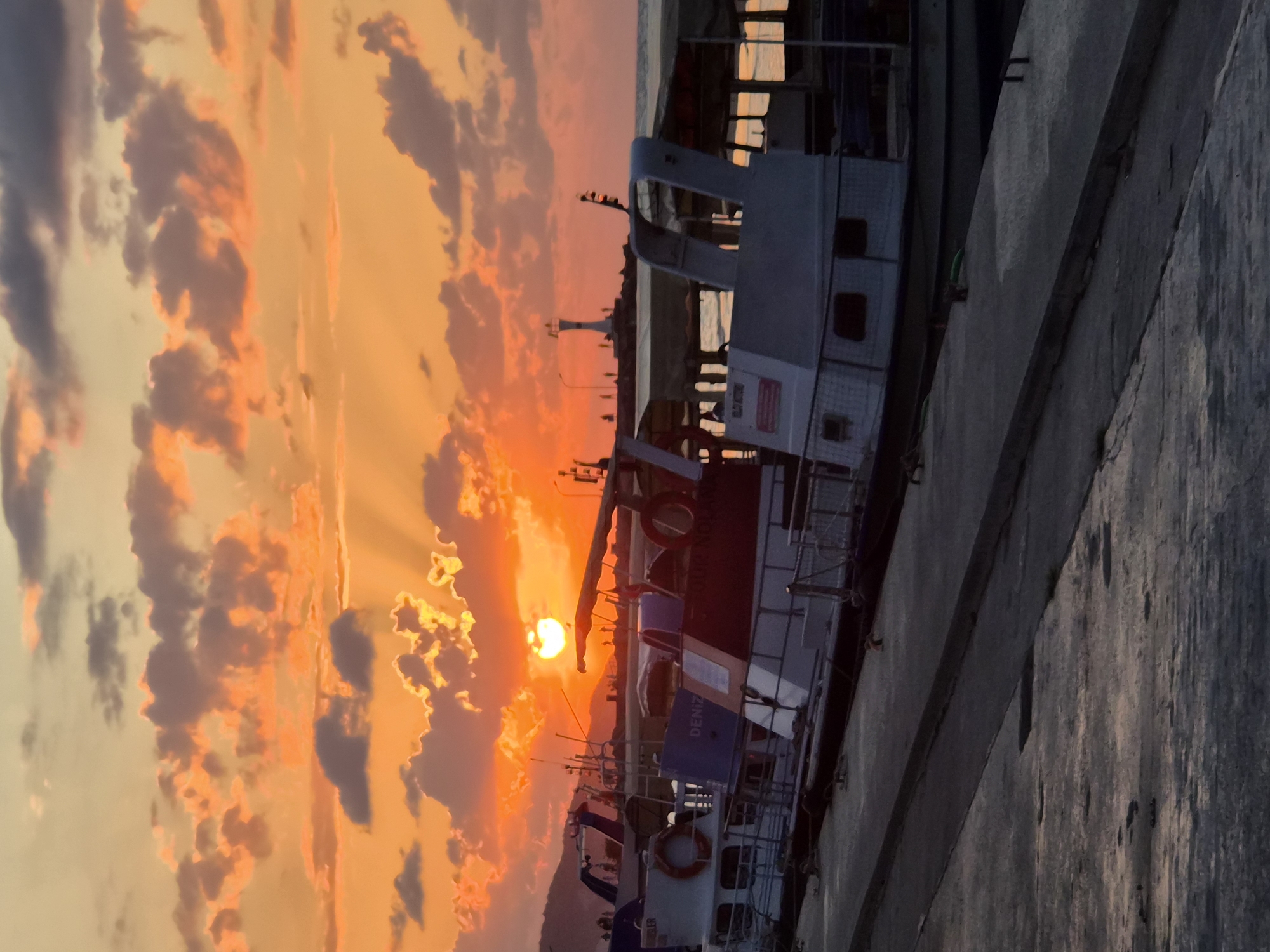The image shows a sunset over the town of Şile, Turkey. The sky is filled with orange and pink clouds, and the sun is setting behind them. In the foreground, there are several boats docked at a pier. In the background, there is a lighthouse. The overall mood of the image is peaceful and serene.