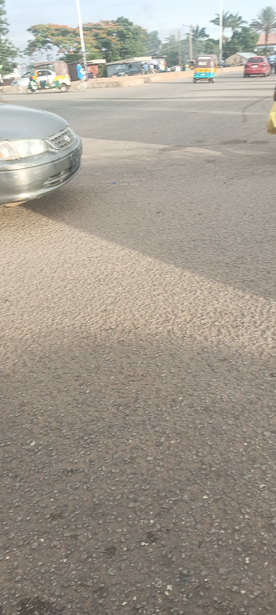 This image presents a detailed street scene in Bukuru, Nigeria, captured during daylight. The foreground is dominated by a grey asphalt road surface exhibiting visible aggregate and minor wear. A distinct, diagonal shadow extends from the lower-left corner, traversing the road surface towards the mid-right.
In the lower-left quadrant, the front bumper and headlights of a silver-grey sedan are partially visible. Moving into the mid-ground, a paved road stretches towards the horizon. On the left side of the street, a person wearing a blue shirt and light-colored trousers is observed walking away from the viewer. Adjacent to this pedestrian, a yellow and white three-wheeled auto-rickshaw (keke Napep) with a brown canopy is stationary or moving slowly on the roadside. Further right, a blue and yellow three-wheeled auto-rickshaw is seen from the rear, traveling away from the viewer. A red sedan is visible in the distance on the right side of the road. On the extreme right edge of the image, a partially visible individual holding a yellow bag is present.
The background features a varied landscape. Numerous trees with green foliage are present, along with at least one tree displaying reddish-orange flowers. Interspersed among the vegetation are several low-rise structures, some with visible corrugated metal roofs, suggesting commercial or residential buildings. A dark blue van is visible behind these structures in the far background. A tall, slender white pole extends vertically from the mid-ground towards the top of the frame. The sky above is light and appears to be hazy or overcast.