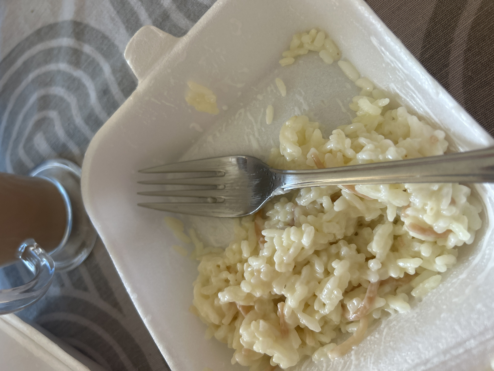 The image shows a close-up view of a white styrofoam container filled with white rice, partially eaten, with a silver fork resting on top of the rice. The rice appears to be creamy and possibly mixed with small pieces of meat. To the left of the container, a portion of a clear glass mug with a brown beverage is visible, suggesting it might be coffee or tea. The background consists of a grey and white striped fabric, possibly a tablecloth or placemat. The lighting suggests it is daytime. The scene is likely a meal being eaten at a table. There is no visible text in the image.