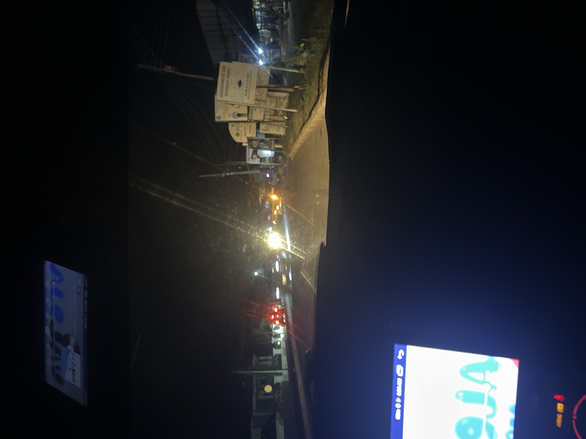 The image is a nighttime view from inside a vehicle, looking forward. The scene depicts a street in Jalingo, Nigeria, illuminated by streetlights and vehicle headlights. On the right side of the road, there are several signs, including one that reads "GOVERNMENT DAY SECONDARY SCHOOL". Further down the street, buildings and shops are visible. Traffic is present, with car taillights and headlights creating streaks of light. The foreground shows the dashboard of the vehicle, with a screen displaying a map or application. The overall atmosphere is that of a typical urban nighttime scene.