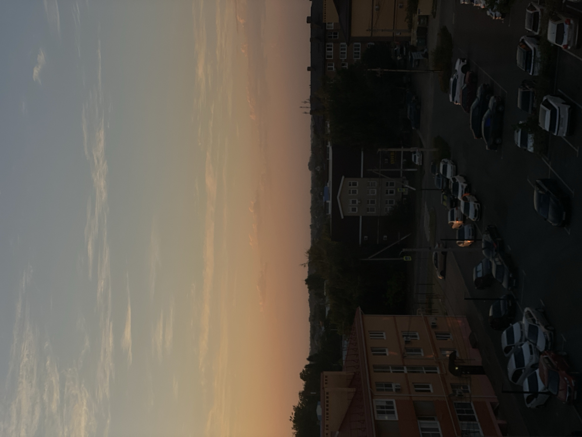The image depicts a dusk scene in Krasnodarskaya Ptitsefabrika, Russia, characterized by a fading sunset casting warm hues across the sky. In the foreground, a multi-story building with an orange facade and a red roof is visible. Adjacent to it, a darker building with visible windows and air conditioning units is present. A parking lot is situated in front of these buildings, filled with numerous parked cars of various models and colors, including white, silver, black, and red vehicles. Further in the background, more buildings and trees are observable, interspersed with what appears to be a wider cityscape or rural landscape under the twilight sky. The sky itself is a gradient of pale blue and soft orange, with scattered thin clouds. No people are visible in the image. The scene conveys a quiet, residential atmosphere at the end of the day.