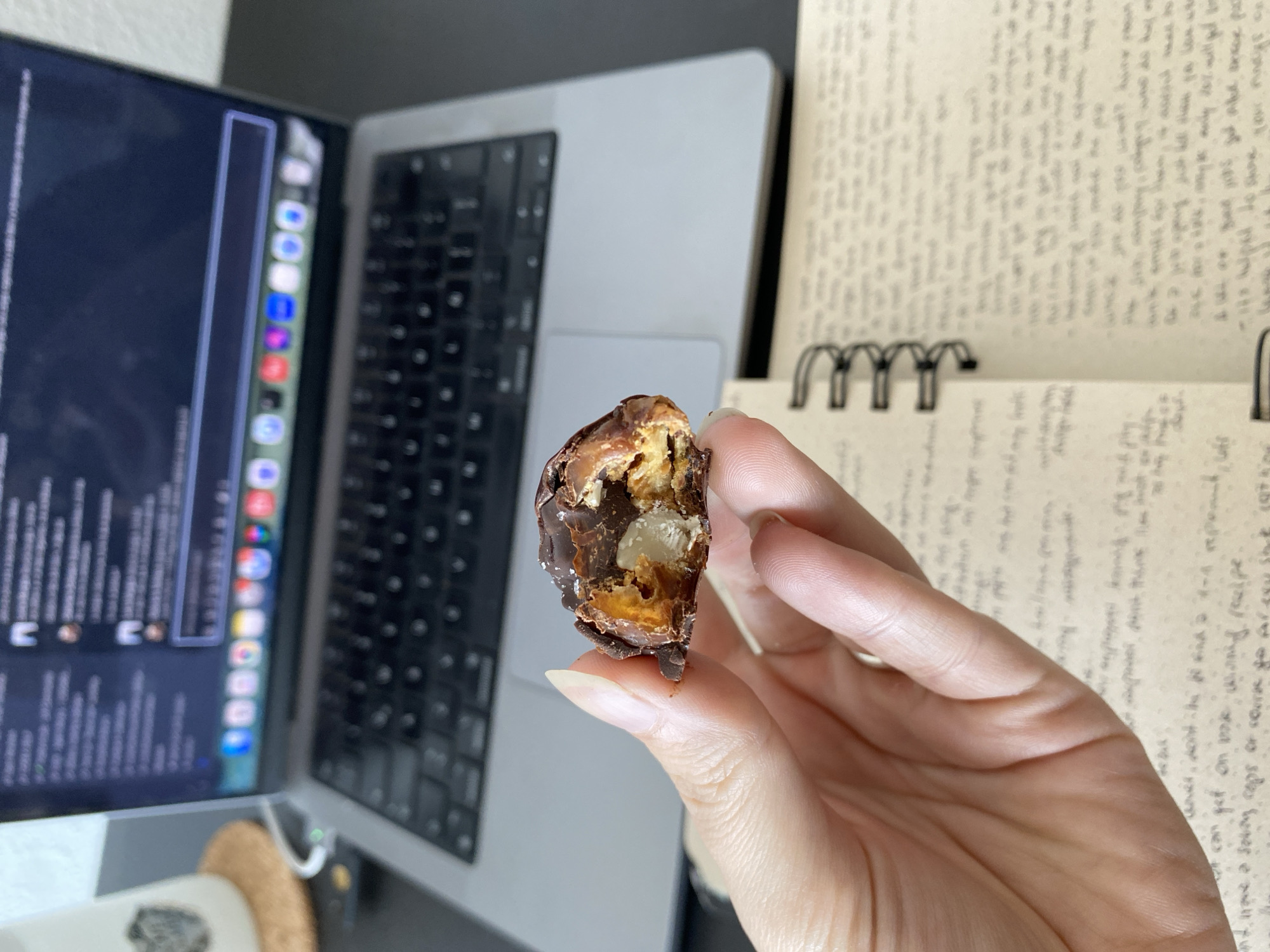 A person is holding a cut date filled with what appears to be an almond or nut and chocolate in their right hand. The hand is fair-skinned and has long nails. Behind the hand, there is a notebook open with handwritten text on its pages. To the left, a laptop computer is open. The laptop's screen displays various icons and text. The overall environment appears to be a desk or workspace. There are no indications of the location being Gijón/Xixón, Spain.