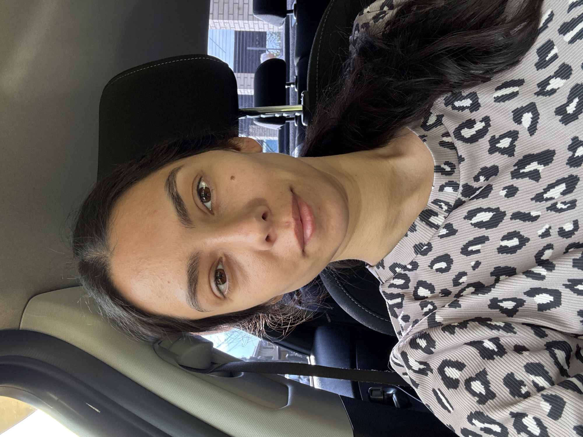 A woman is sitting in the driver's seat of a car, looking at the camera. The car appears to be parked, with the interior visible. The woman has long, dark hair pulled back into a ponytail and is wearing a grey top with a black and white leopard print. The background shows the interior of the car and through the window, glimpses of buildings and trees, suggesting an urban or suburban setting. There is no indication of the specific location within Chaikovsky, Russia.