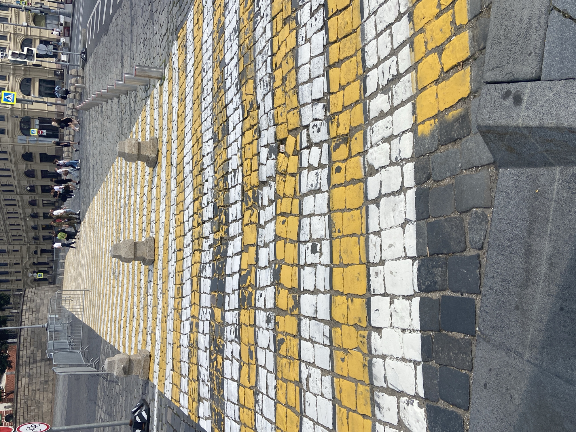 The image captures a pedestrian crossing on a cobblestone street in Moscow, Russia. The crossing is marked with alternating yellow and white stripes, painted onto the uneven cobblestones, with some areas showing wear and tear on the paint. Two stone bollards are positioned at the near side of the crossing, one on each side, acting as barriers. In the background, several people are walking across the street, some in casual clothing and others in what appear to be military uniforms. A yellow pedestrian crossing sign is visible on a pole in the middle ground, along with traffic lights and the facades of buildings lining the street. The environment suggests a public space, possibly a square or a busy intersection, with a mix of modern infrastructure and historic street surfaces.