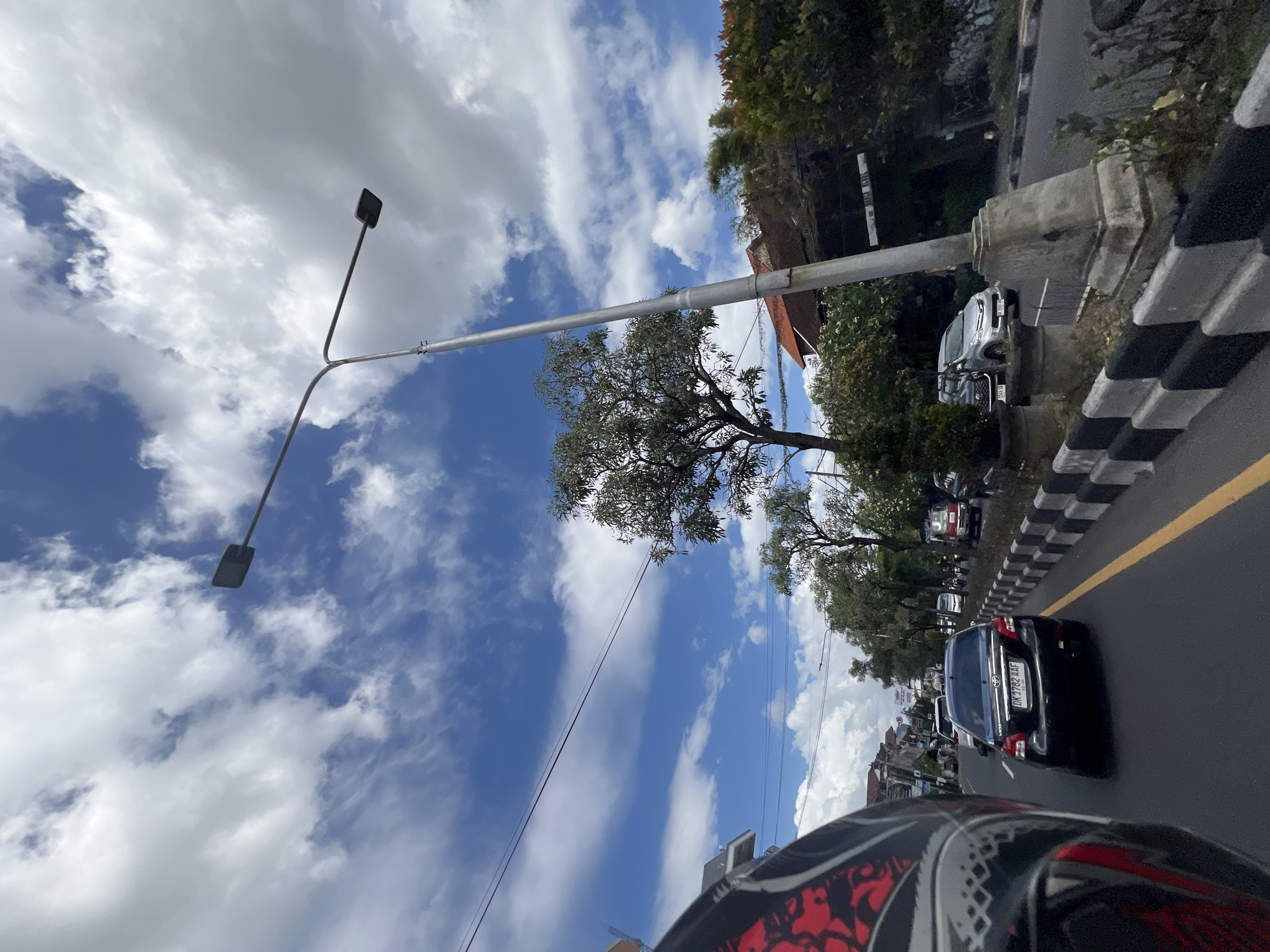 The image shows a street scene in Denpasar, Indonesia, on a cloudy day. The perspective is from a motorcycle, with the rider's helmet visible in the foreground left. The street is lined with trees and buildings, and there is moderate traffic, including cars and motorcycles. A tall street light stands prominently in the center of the frame. The sky is a mix of blue and white clouds, and overhead power lines are visible. The road has a yellow dividing line and a concrete barrier with black and white markings. The overall impression is of a typical urban street in a tropical climate.