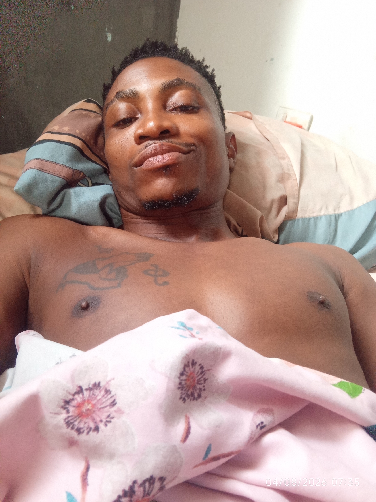A person is lying on a bed, partially covered by a pink floral sheet. Their chest and face are visible. The person has a tattoo on their chest. A pillow with striped fabric is behind their head. The environment appears to be indoors, possibly a bedroom. A timestamp in the lower right corner of the image reads "04/08/2026 07:35".