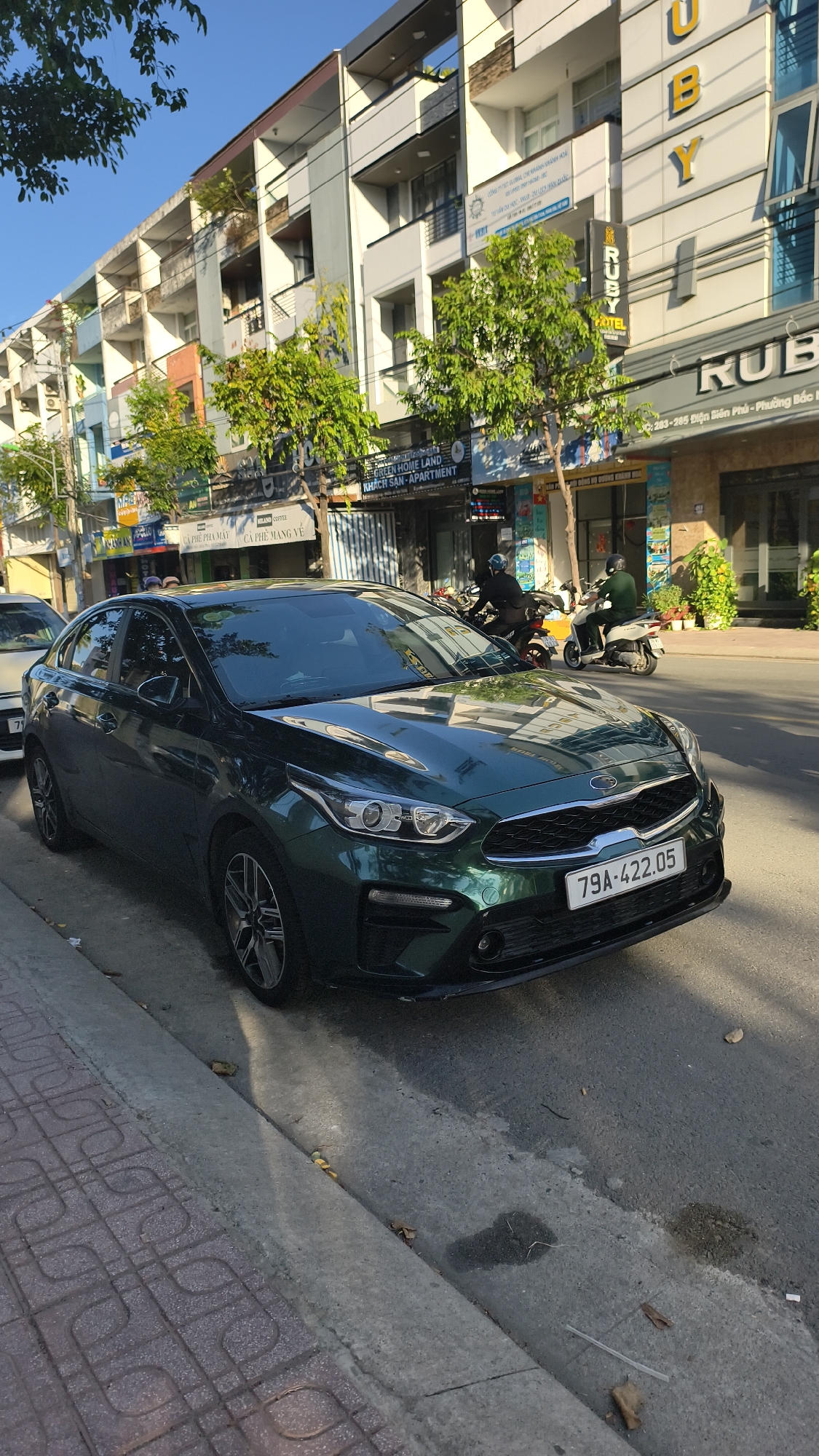 A dark green Kia sedan is parked on the right side of a street in Nha Trang, Vietnam. The car faces towards the viewer's right and is positioned at an angle. Its license plate reads "79A-422.05". The vehicle is parked alongside a sidewalk with a patterned tile surface and a concrete curb. The street is paved with asphalt and appears to be a city street with buildings lining the opposite side. These buildings are multi-story structures with various storefronts on the ground floor. Trees with green foliage are visible along the street. In the background, two individuals on motorbikes are traveling down the street. One motorbike is white, and the other is a darker color. The sky above is clear and blue, indicating a sunny day. The overall scene depicts a typical urban street in Nha Trang.