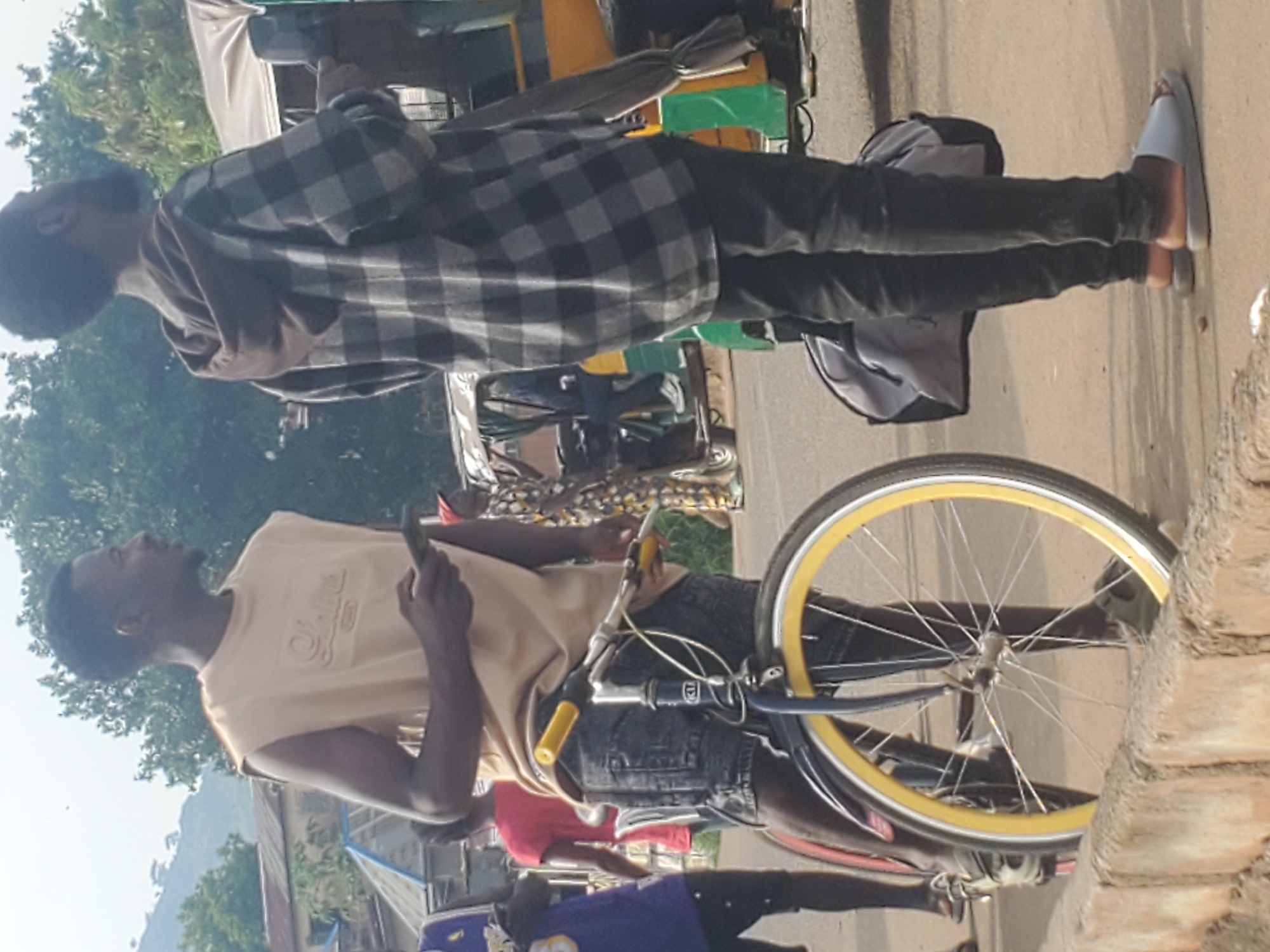 In Gwong, Nigeria, a street scene unfolds with two young men prominently featured. The man on the left, wearing a sleeveless tan t-shirt and dark denim shorts, stands next to a bicycle with a yellow rim. He holds a mobile phone in his right hand. To his right, another man, clad in a grey and black plaid hooded shirt and dark trousers, looks towards the right of the frame. Behind them, a three-wheeled auto-rickshaw, a common mode of transportation in Nigeria, is visible, with a person seated in the driver's position. The backdrop consists of trees and buildings, suggesting an urban or semi-urban environment. The ground is paved, and a low brick wall runs along the foreground. The overall lighting indicates it is daytime.