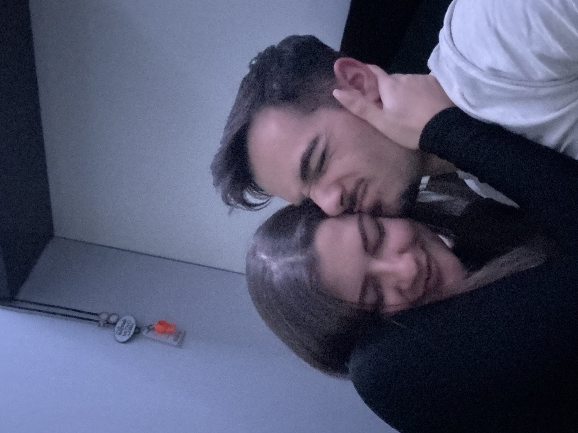 The image depicts two individuals, an adult female and an adult male, in close physical proximity within an indoor setting. The female, positioned on the left, has dark hair parted centrally and is wearing a dark long-sleeved top. Her face is turned towards the male, with eyes closed or downcast and a subtle smile. Her right hand is visible resting on the male's left jawline. The male, on the right, has dark hair and facial stubble, wearing a light-colored t-shirt. His face is directed towards the female, displaying a scrunched expression with squinted eyes and a slightly open mouth. The background consists of light blue-grey walls. In the upper left corner, a lanyard hangs, from which a white oval badge inscribed with "Sofia Tech Park" and "MITKO" is visible, along with an orange rectangular object contained within a clear plastic holder. This identifies the location as Sofia Tech Park in Sofia, Bulgaria. The scene is illuminated by dim, cool-toned ambient light.