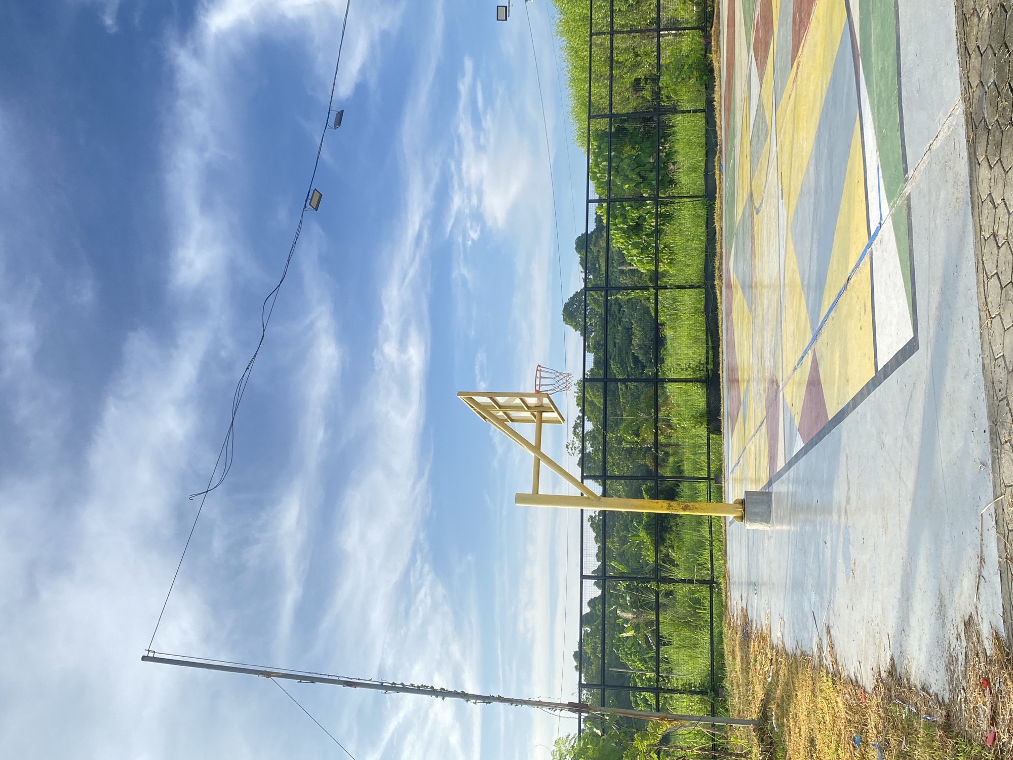 An outdoor basketball court is depicted under a partly cloudy blue sky. The court surface is concrete and painted with a geometric pattern of yellow, green, red, and white shapes. A yellow basketball hoop with a red net stands on the court. In the background, a black metal fence encloses the court, beyond which lies lush green vegetation and trees. Two square floodlights are suspended from wires strung across the sky. A metal pole with attached wiring stands to the left of the basketball hoop. The foreground shows a patch of dry grass and debris. The scene appears to be in a recreational area within Davao City, Philippines, characterized by its tropical foliage and bright, open sky.