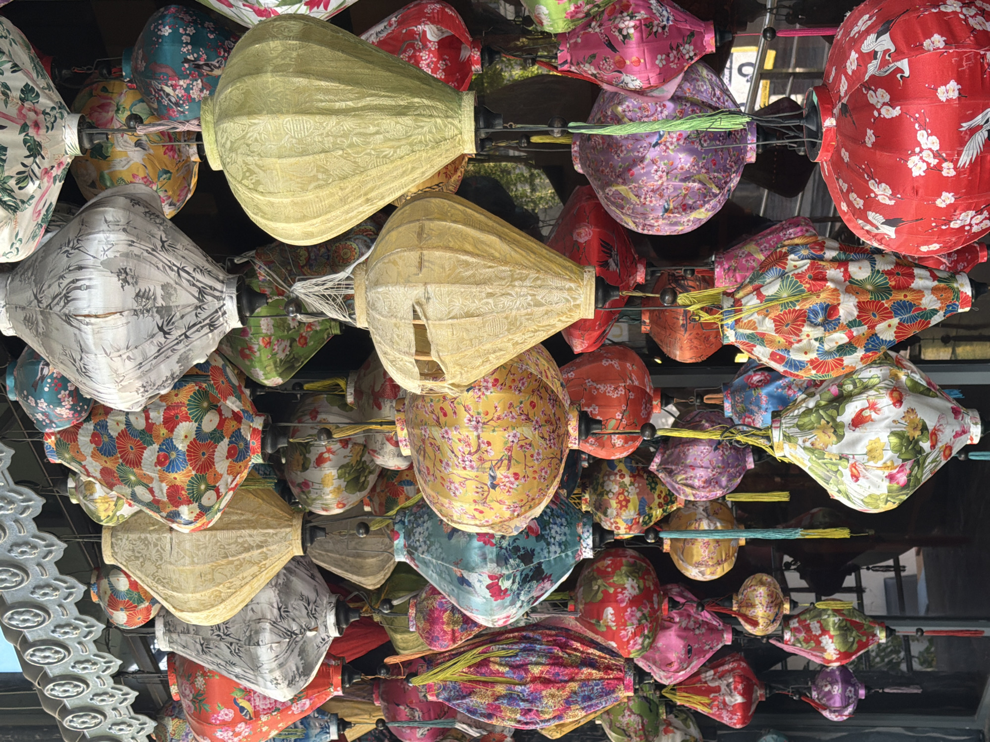 The image shows a vibrant display of numerous colorful silk lanterns hanging outdoors, likely in a market or along a street in Nha Trang, Vietnam. The lanterns are of various shapes and sizes, many adorned with intricate floral patterns, birds, and other traditional Asian motifs. Some are cylindrical, others diamond-shaped or bulbous. They are illuminated by natural light, casting shadows and creating a visually rich and textured scene. The background is blurred but suggests an urban environment with some architectural elements, possibly a shop front or a decorative facade above the lanterns. There are no people visible in the frame. The lanterns are suspended by strings, some with decorative tassels.