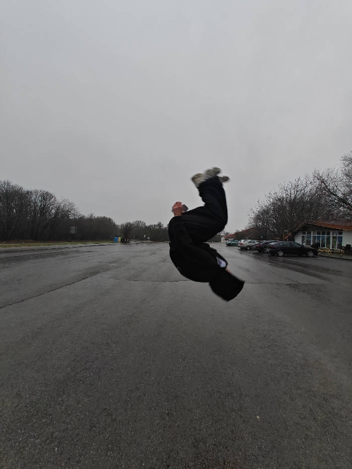 The image captures an individual in mid-air performing an acrobatic flip on a wet asphalt surface in Shumen, Bulgaria, under an overcast sky. The subject is dressed in dark clothing, possibly a tracksuit, and light-colored athletic shoes, exhibiting motion blur around the feet and head due to movement. The ground is dark asphalt, visibly wet from rain, reflecting the dull light. In the background, a dense line of bare deciduous trees runs along the horizon on the left. To the right, a single-story building with a light-colored facade and a reddish-brown tiled roof is visible, fronted by an asphalt parking lot. Several vehicles, including a dark sedan and a silver hatchback, are parked in front of the building. Further left in the background, near the tree line, two large rectangular structures and a blue and yellow recycling bin are discernible.