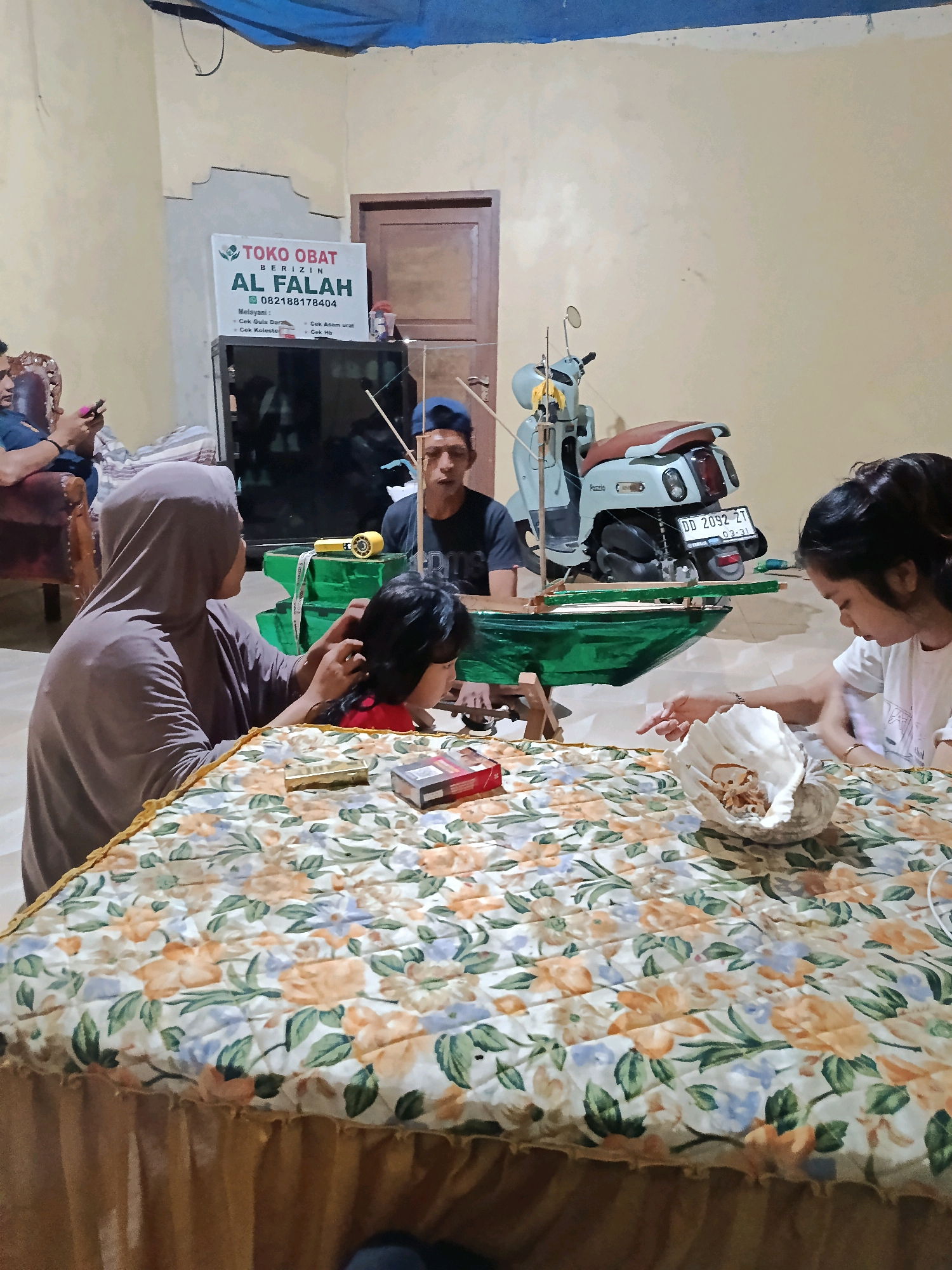 In an indoor setting, likely a home in Bulukumba, Indonesia, a woman wearing a hijab is braiding the hair of a young girl sitting opposite her at a floral-patterned table. The girl is wearing a red shirt. A man wearing a blue cap and dark shirt is visible in the background, looking at something on the table. To the right of the man, a light blue and brown scooter is parked. A model boat, painted green, is positioned on the table between the individuals. On the table, a large seashell contains small, light-colored items, possibly food. A pack of cigarettes and a lighter are also on the table, near the woman and child. In the background, a sign for "TOKO OBAT AL FALAH" (Al Falah Medicine Shop) is visible on the wall, along with a television and a door. The lighting in the room appears to be artificial.