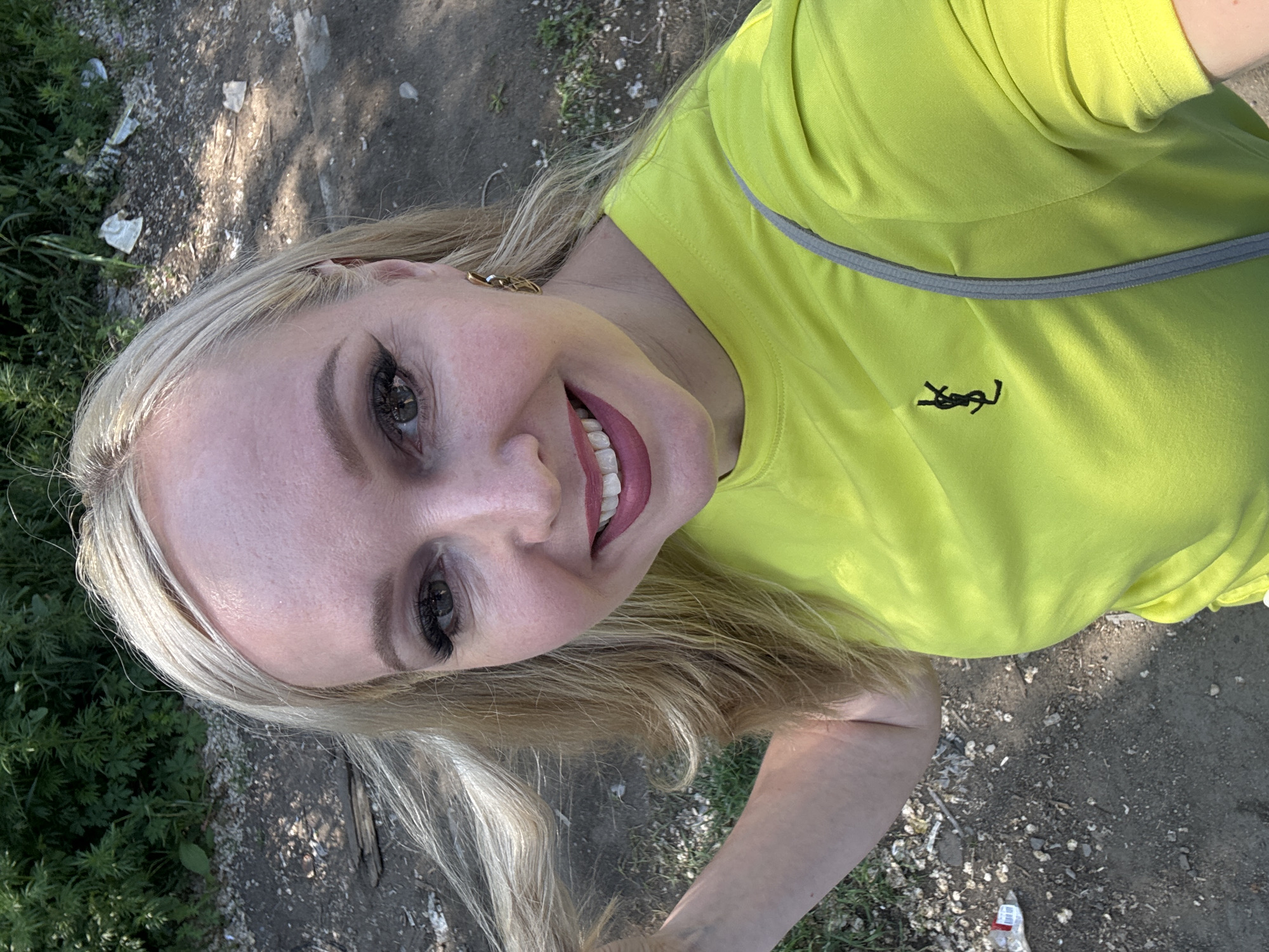 The image shows a woman with blonde hair and fair skin, wearing a bright yellow t-shirt with a black logo. She is outdoors, with greenery and dirt in the background. The lighting suggests it is daytime. The specific location in Komsomolsk-on-Amur, Russia, cannot be determined from the visual information provided.