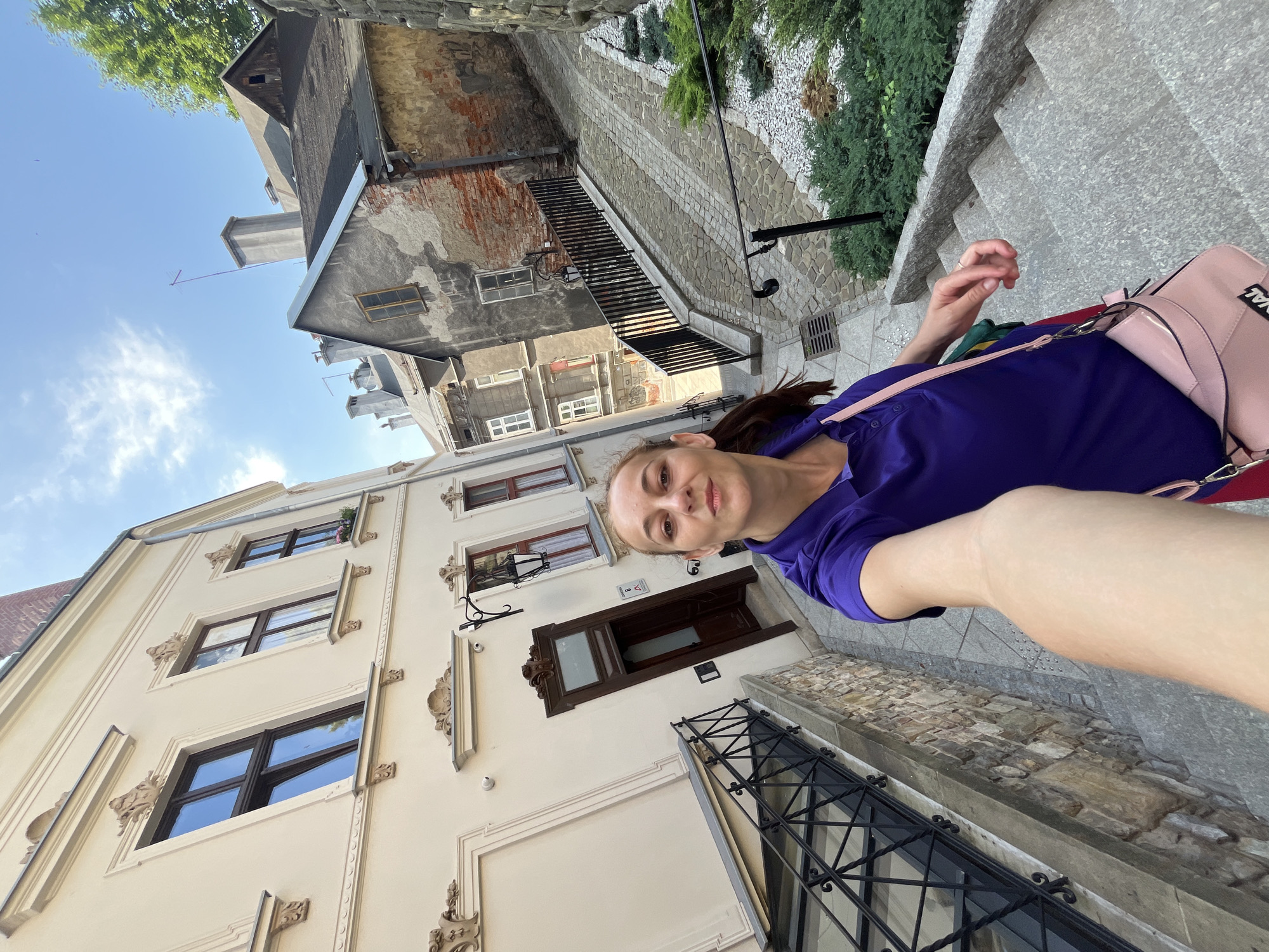 A woman in a purple polo shirt and a pink crossbody bag is taking a selfie while standing on a stone staircase in a narrow street. She is looking at the camera with a neutral expression. The street is lined with old buildings on both sides, with cobblestone pavement and a metal railing along the staircase. The buildings have a mix of architectural styles, with some appearing to be renovated and others showing signs of age. The sky is blue with some scattered clouds. The overall scene suggests an urban environment with historical architecture.