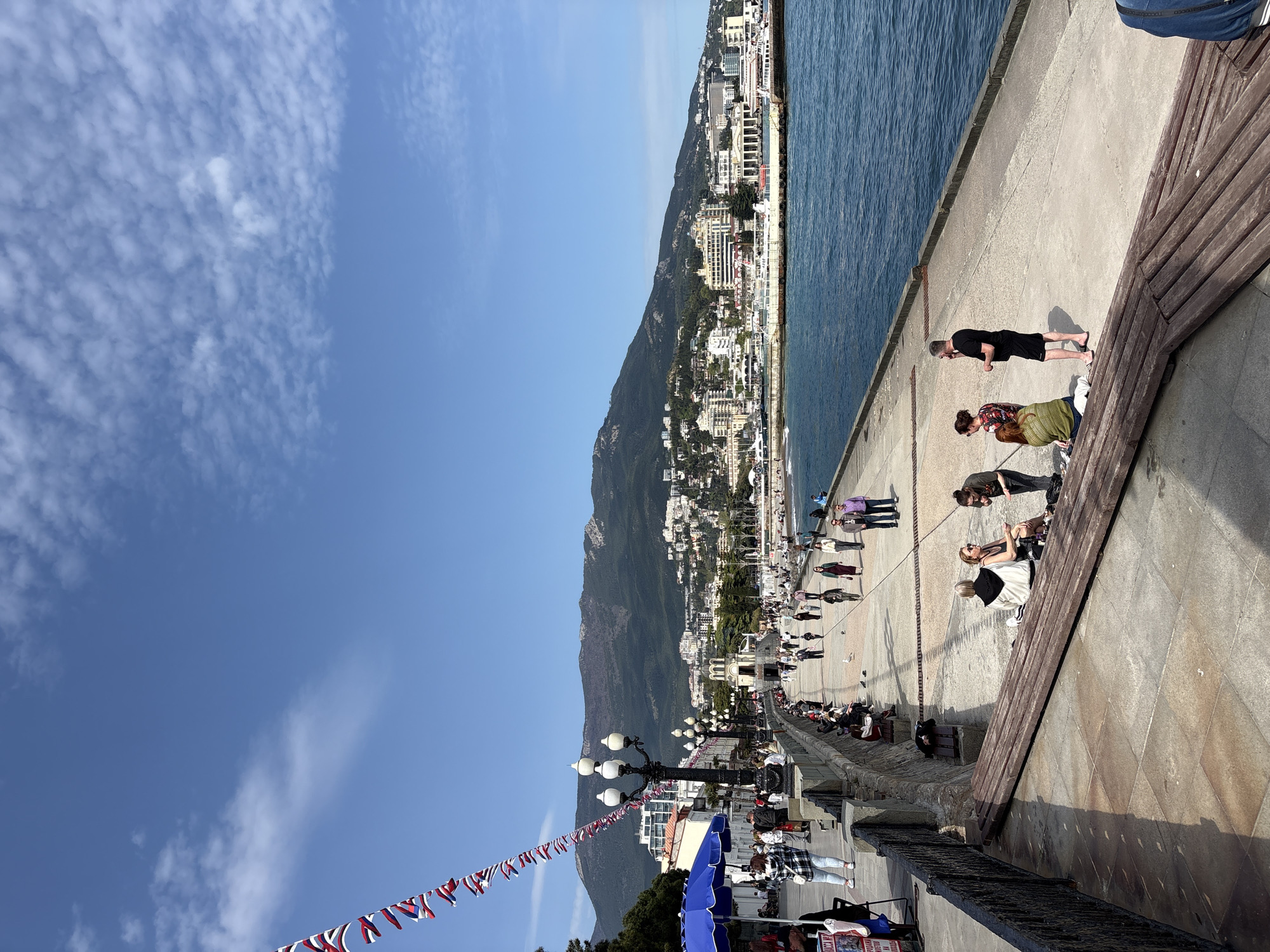 The image depicts a crowded scene on the city promenade in Ялта, Ukraine, adjacent to the coastline. Several people are walking along the promenade, while others are sitting on benches along the edge. One person is standing near the edge, looking out at the water. The backdrop features a mountain and a series of buildings situated along the mountain's slope. Above, the sky is blue with scattered clouds. Decorations, in the form of strung flags, are visible along the street, and light posts are visible.